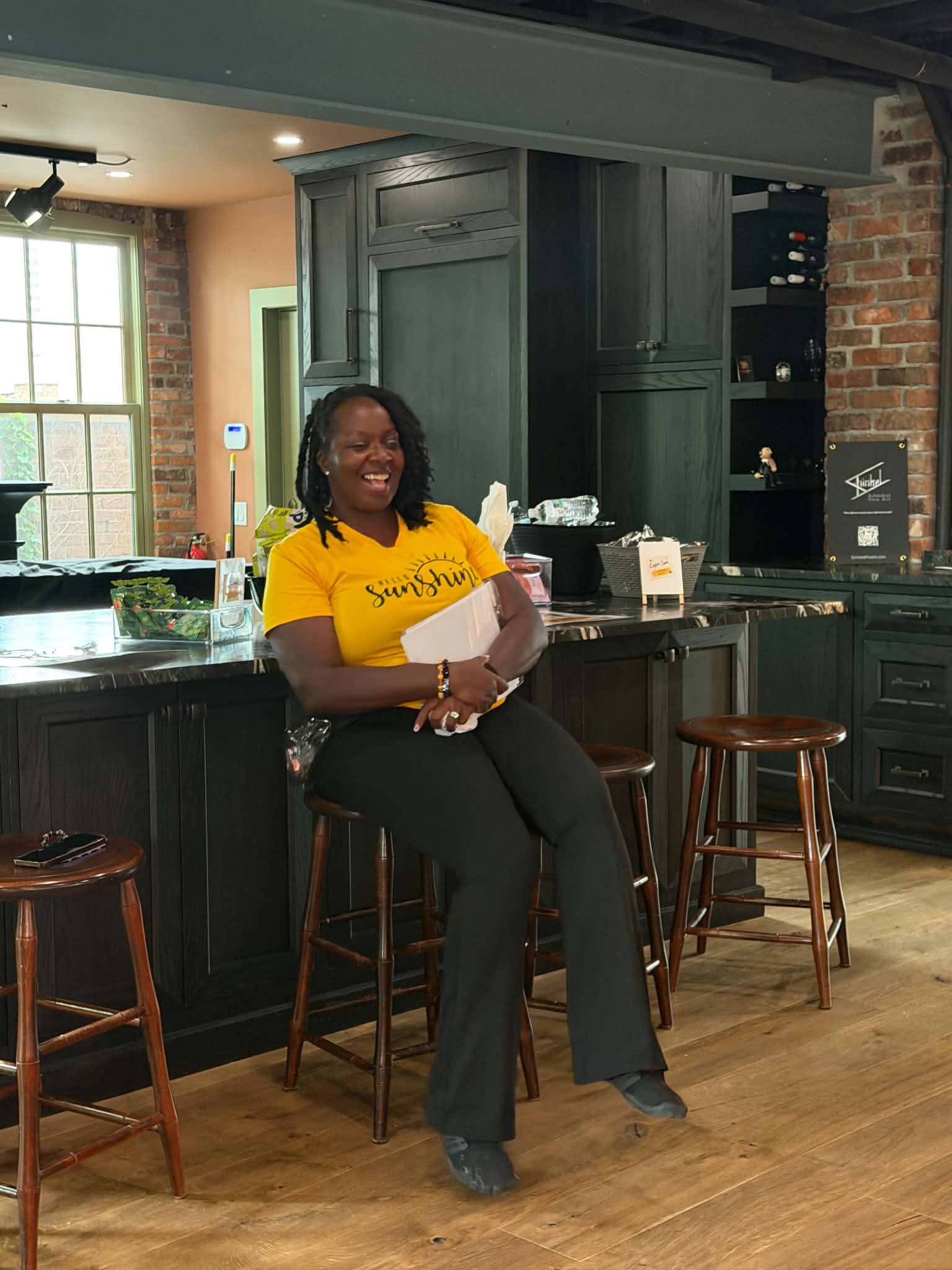 Woman sitting on a barstool in a kitchen, smiling, wearing a yellow shirt and black pants, with green cabinets and brick wall behind her.