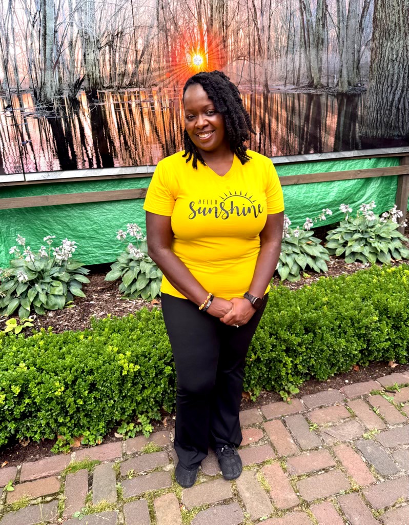 Woman with curly hair smiling, wearing a yellow shirt and black pants, standing outdoors near greenery and a pond at sunset.