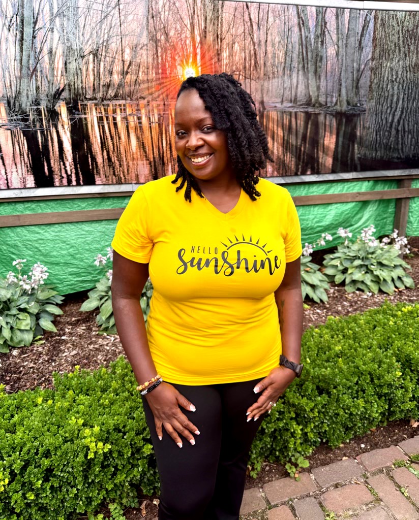 Smiling woman in yellow shirt with 'hello sunshine' text, standing outdoors with greenery and a mural background.