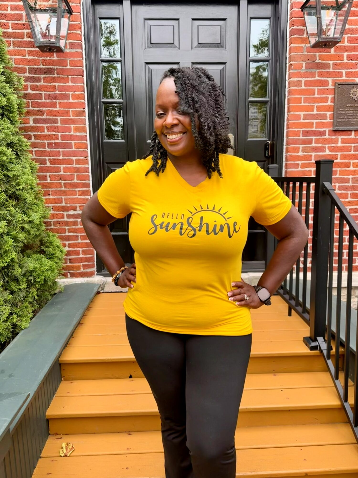 Smiling woman in yellow shirt with 'Sunshine' text, standing on front porch steps, brick house background, black pants, hands on hips.