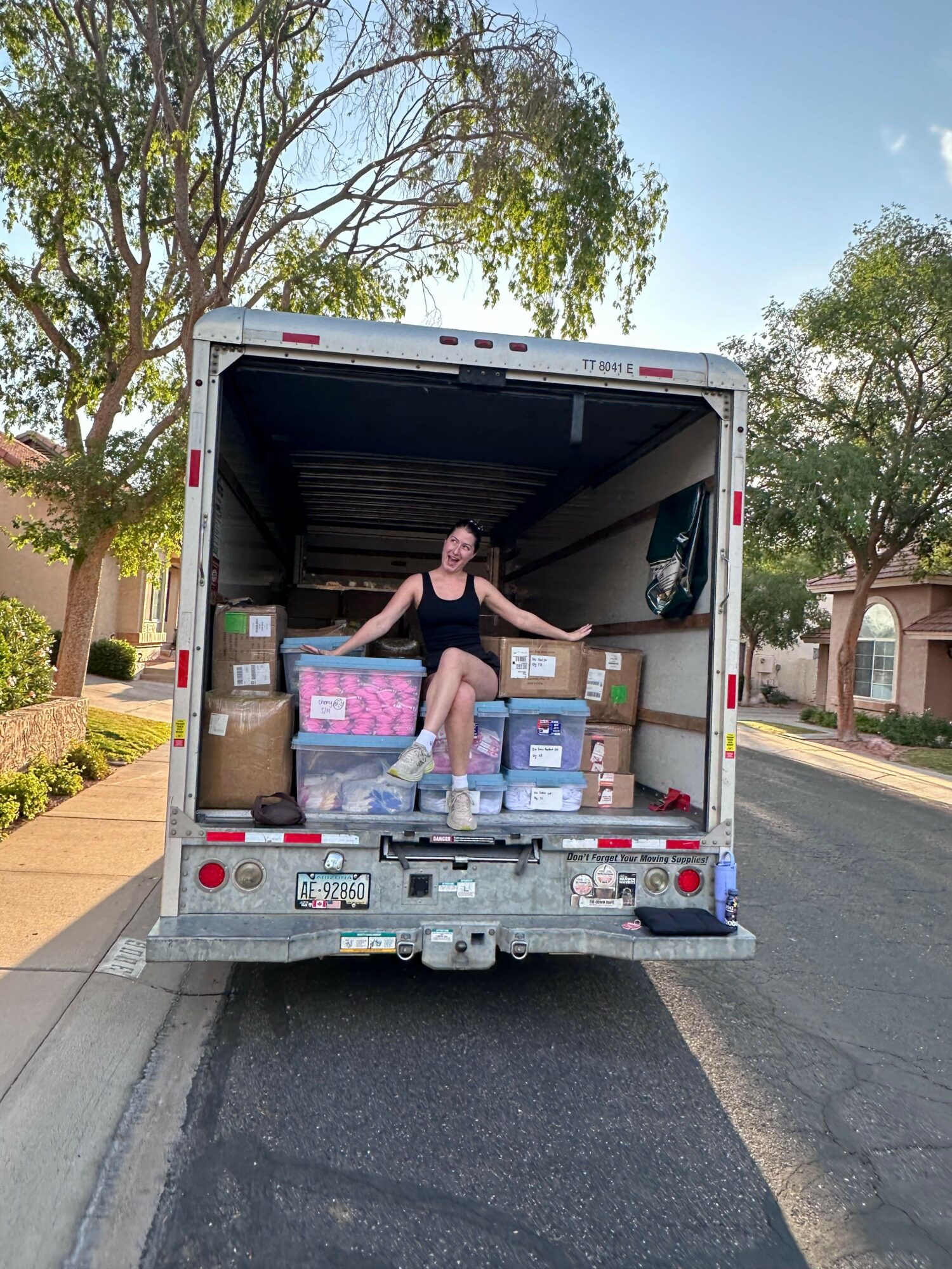 Person loading boxes and supplies into a truck parked on a street with trees and buildings nearby.