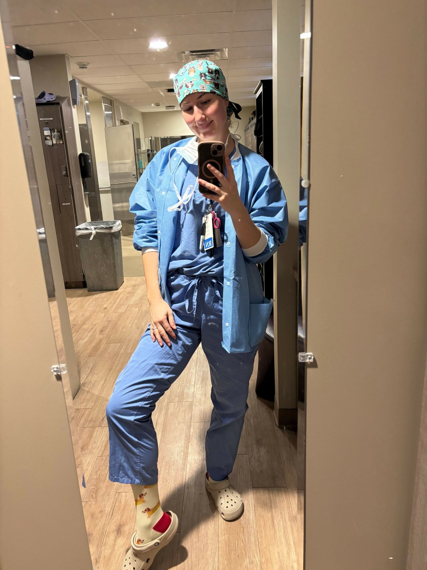Person taking a mirror selfie in a bathroom, wearing a blue jacket, blue pants, and a colorful hat, with a hand on their hip.