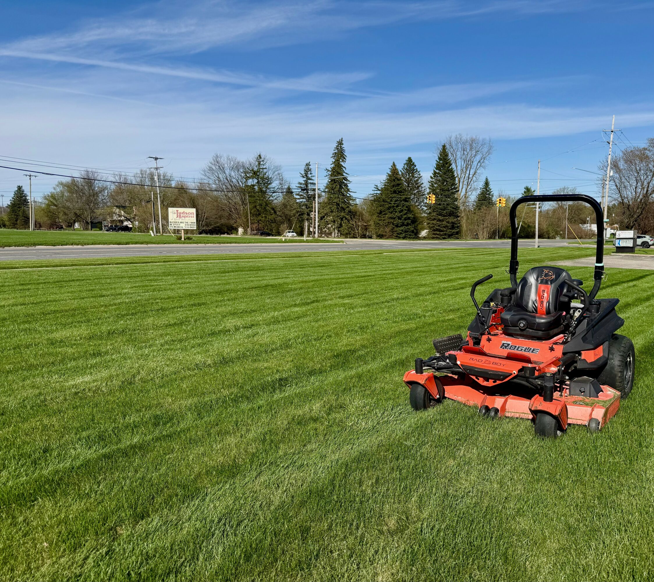 Lawn mower on a grassy field with trees, road, and blue sky in the background.