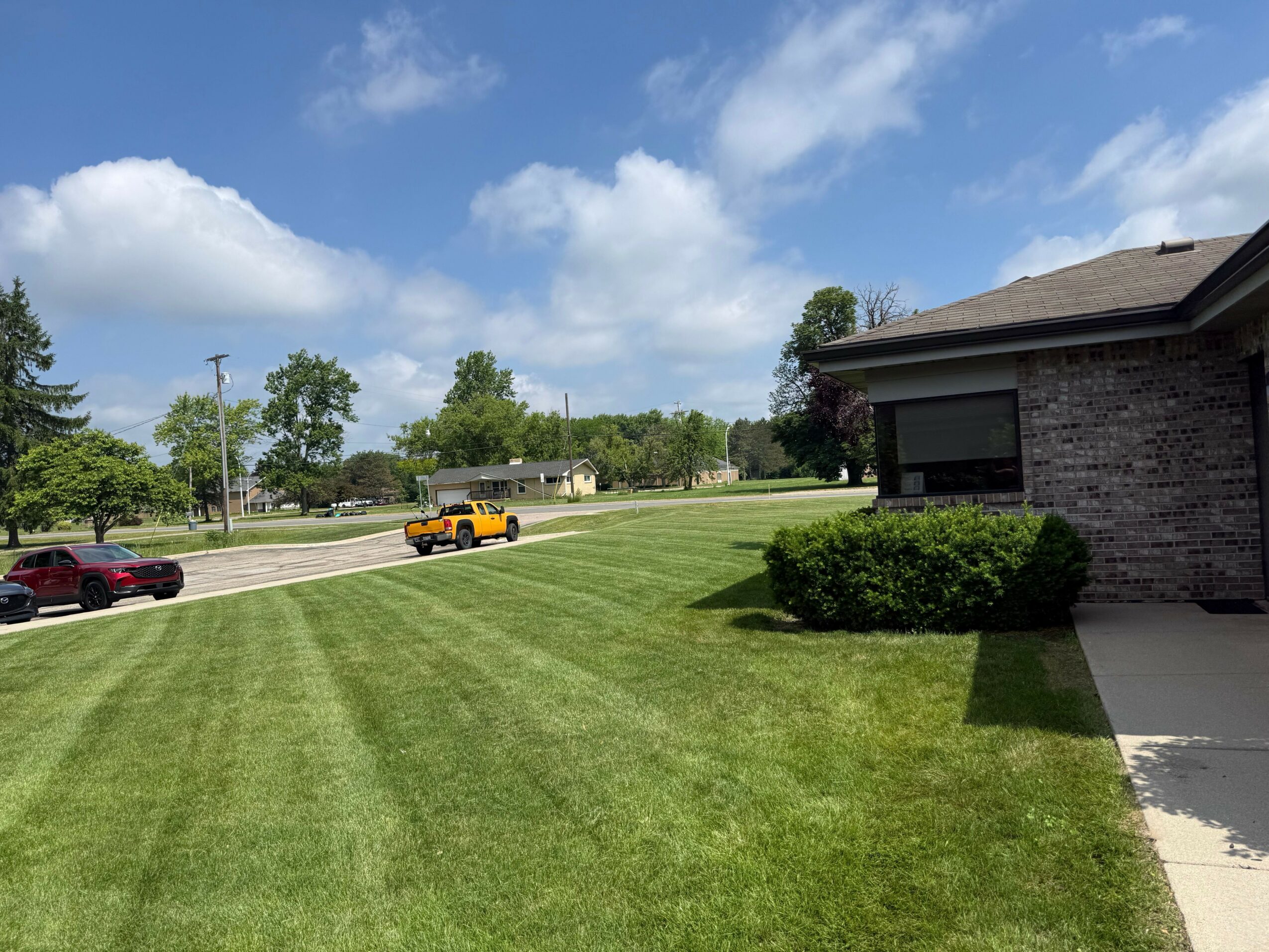 A grassy area with a sidewalk, parked cars, and trees under a partly cloudy sky.