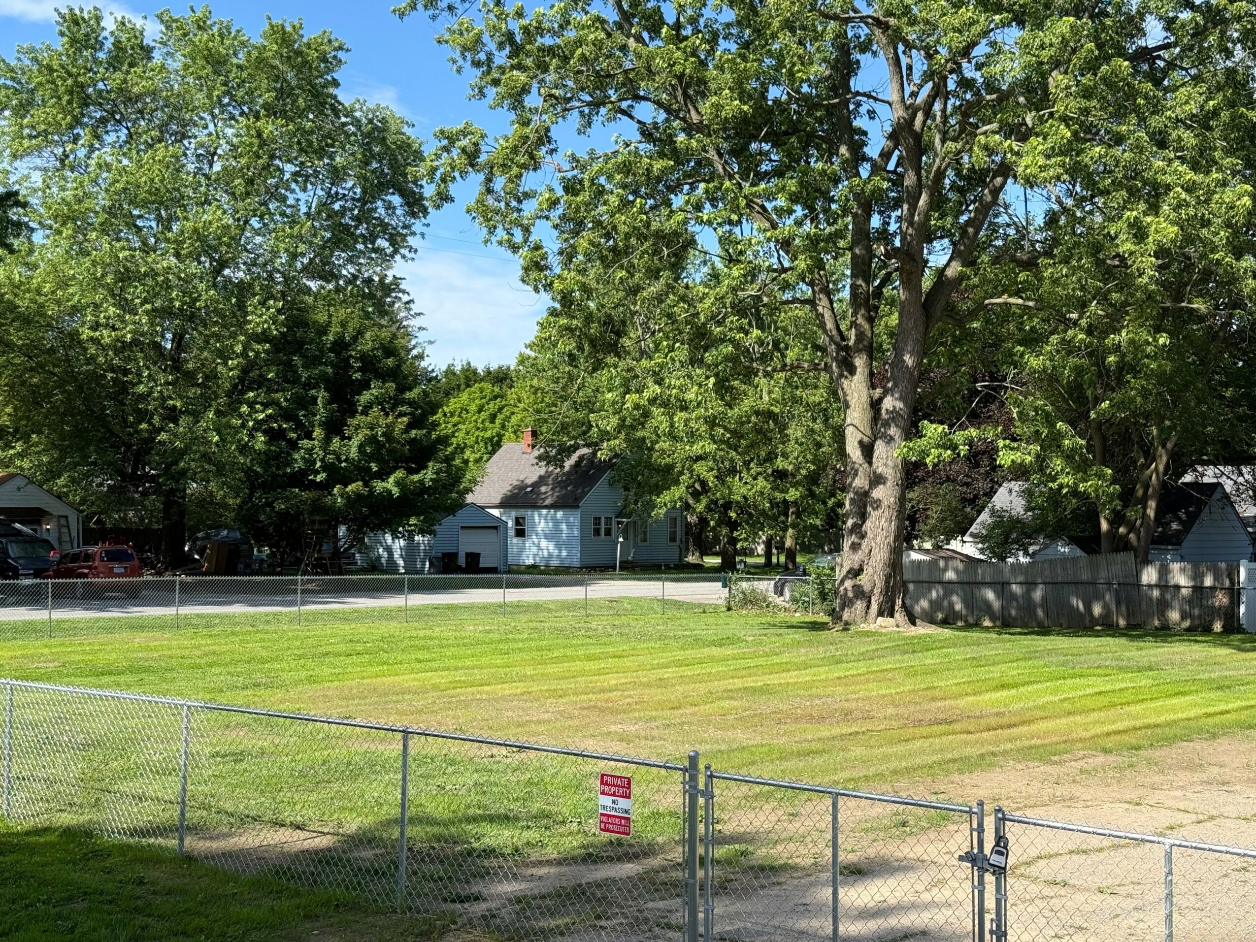 Open grassy field with trees and houses in background, fenced area with a small sign, sunny day with blue sky.