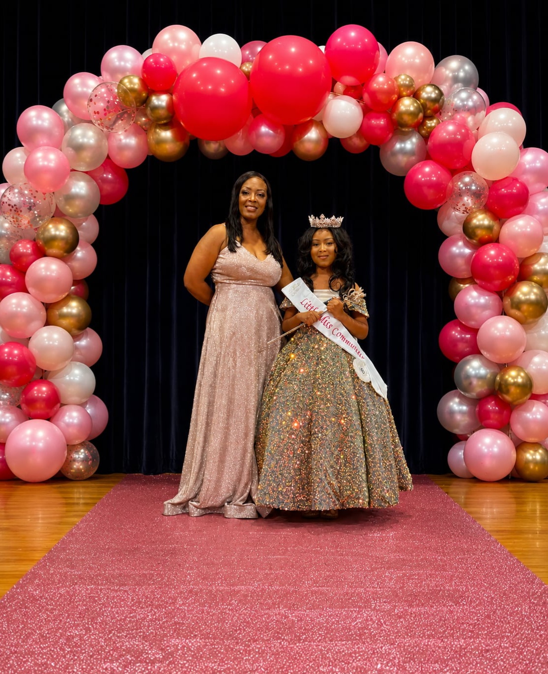 Two women stand under a balloon arch, one wearing a crown and sash, the other in a long dress, on a red carpet.