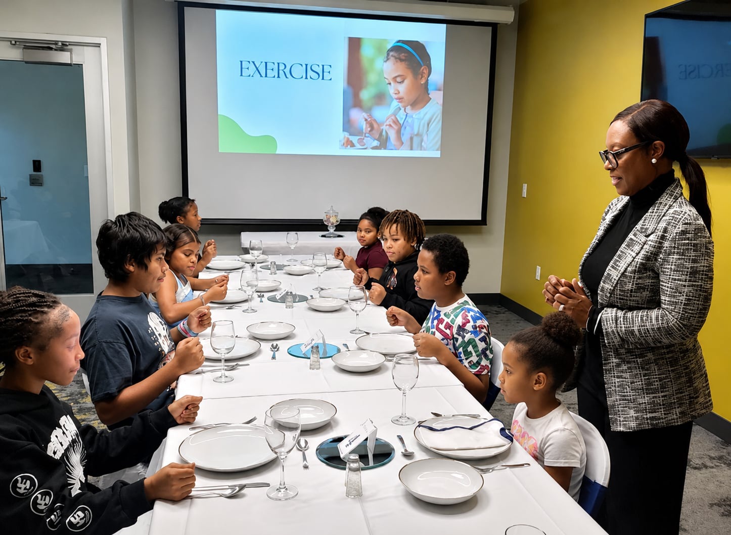 Children seated at a table with plates, listening to a woman standing nearby in a room with a presentation screen.