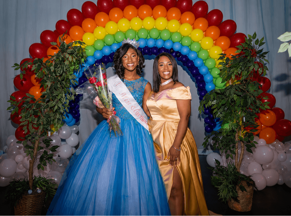 Two women in formal dresses stand in front of a rainbow balloon arch with greenery, one holding flowers, celebrating a special event.