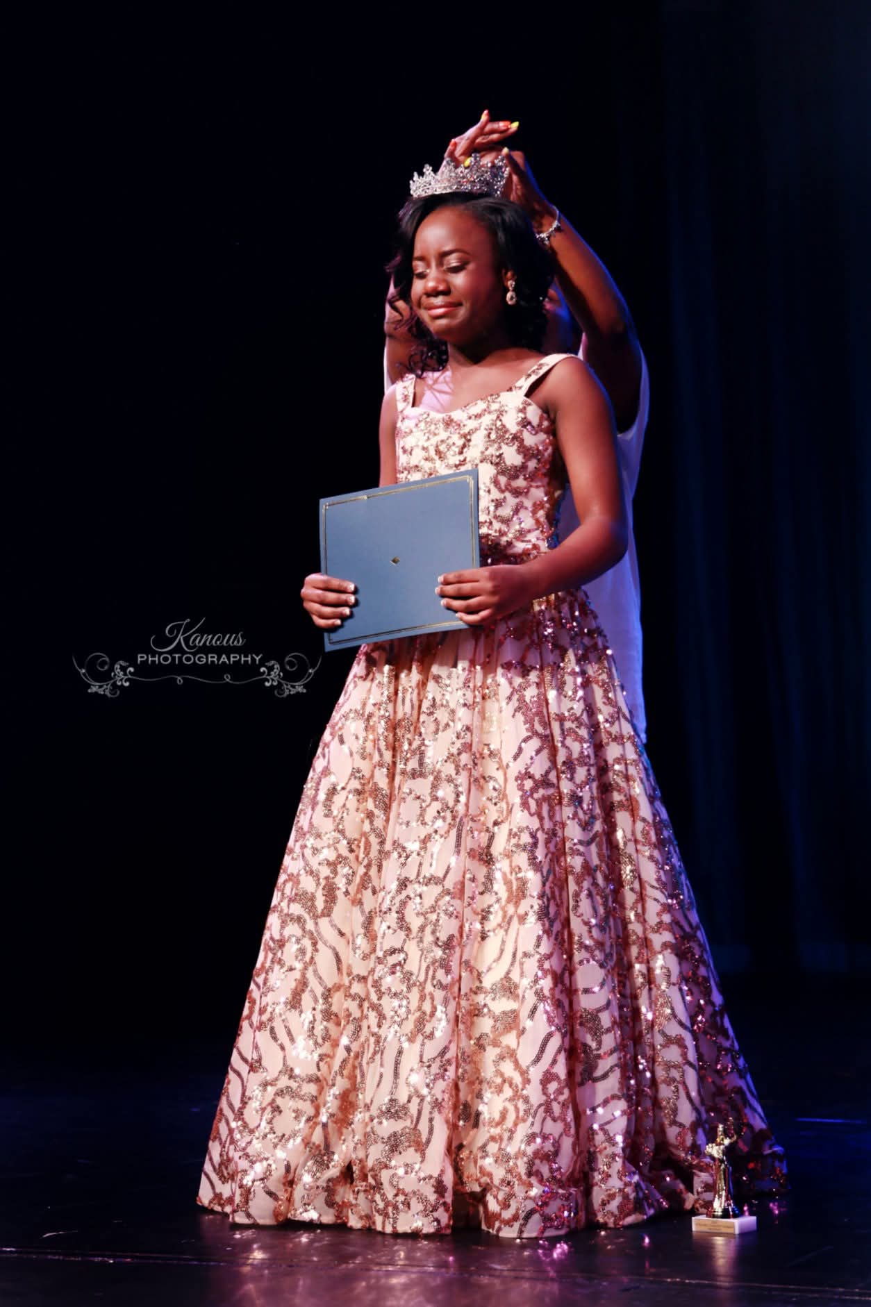 Young girl in a pink and gold gown holding a certificate, standing on stage with a dark background.