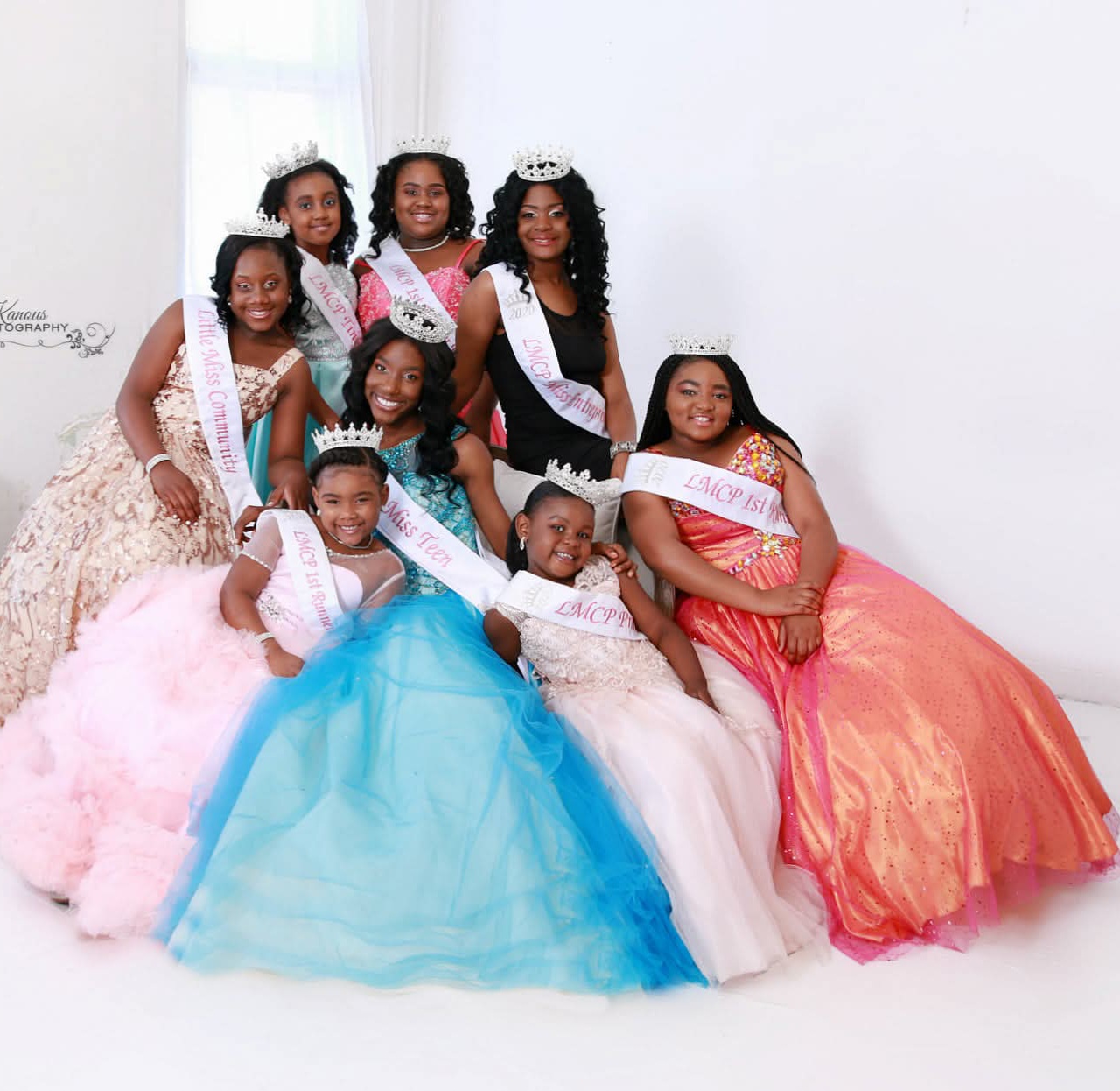 Group of young girls in colorful dresses and tiaras, wearing sashes, posing indoors against a white background.