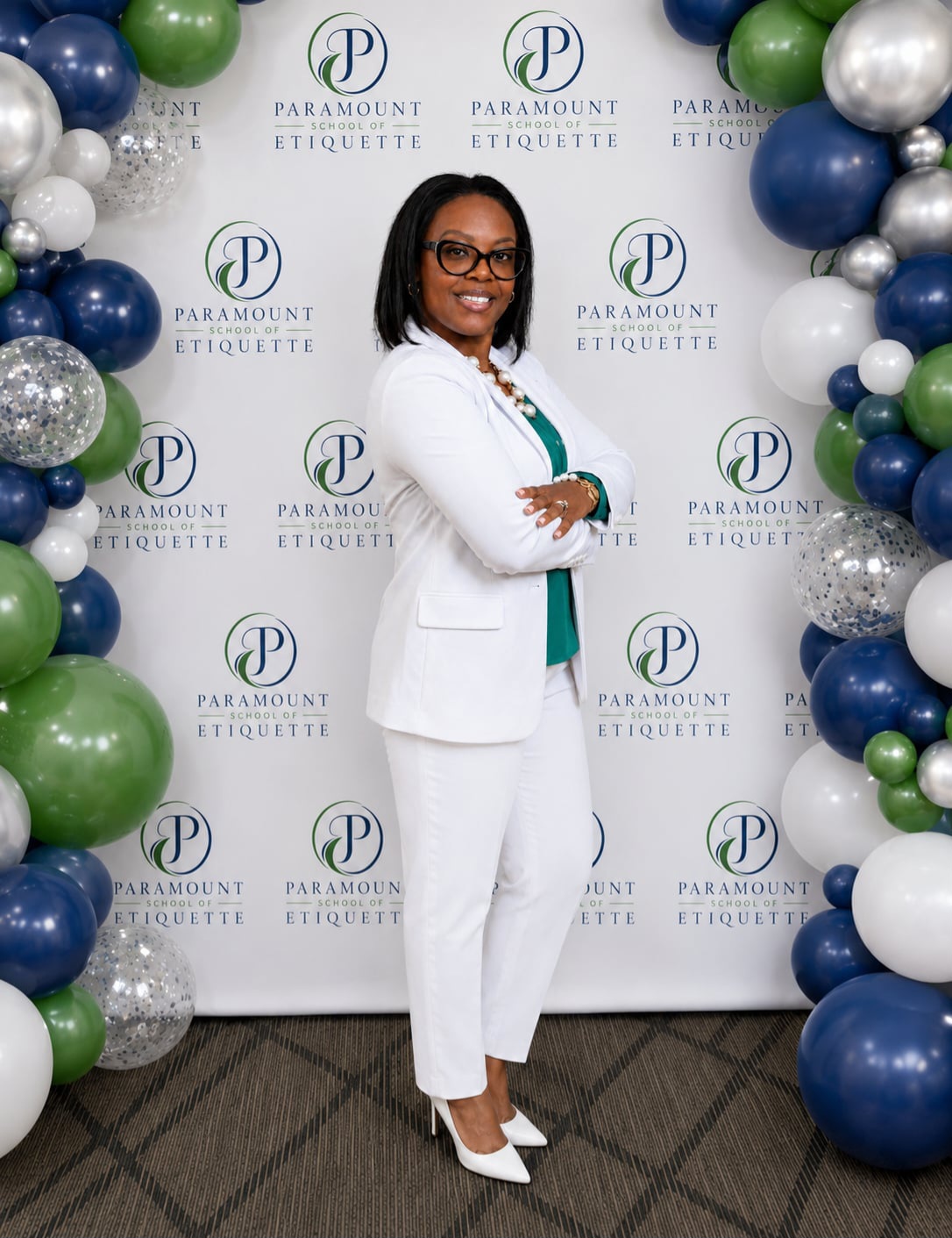 Woman in white suit standing with arms crossed, smiling, surrounded by balloon arch, backdrop with Paramount Etiquette logo.