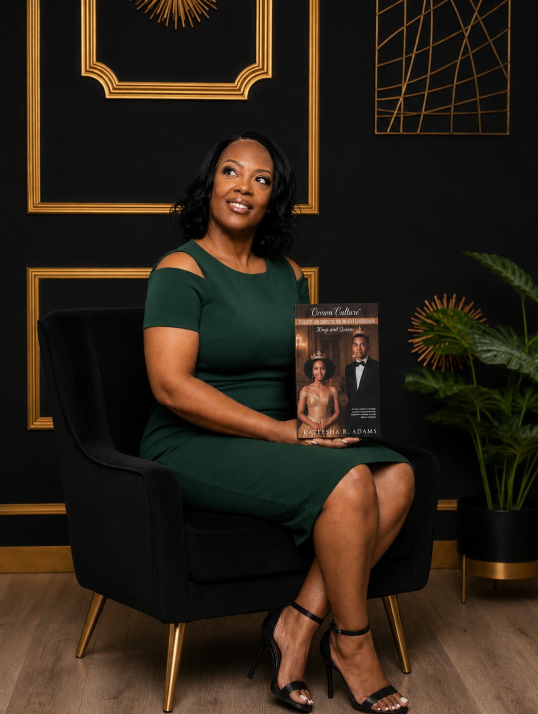 Woman in green dress sitting on black chair holding a magazine, with black and gold wall decor and a plant nearby.