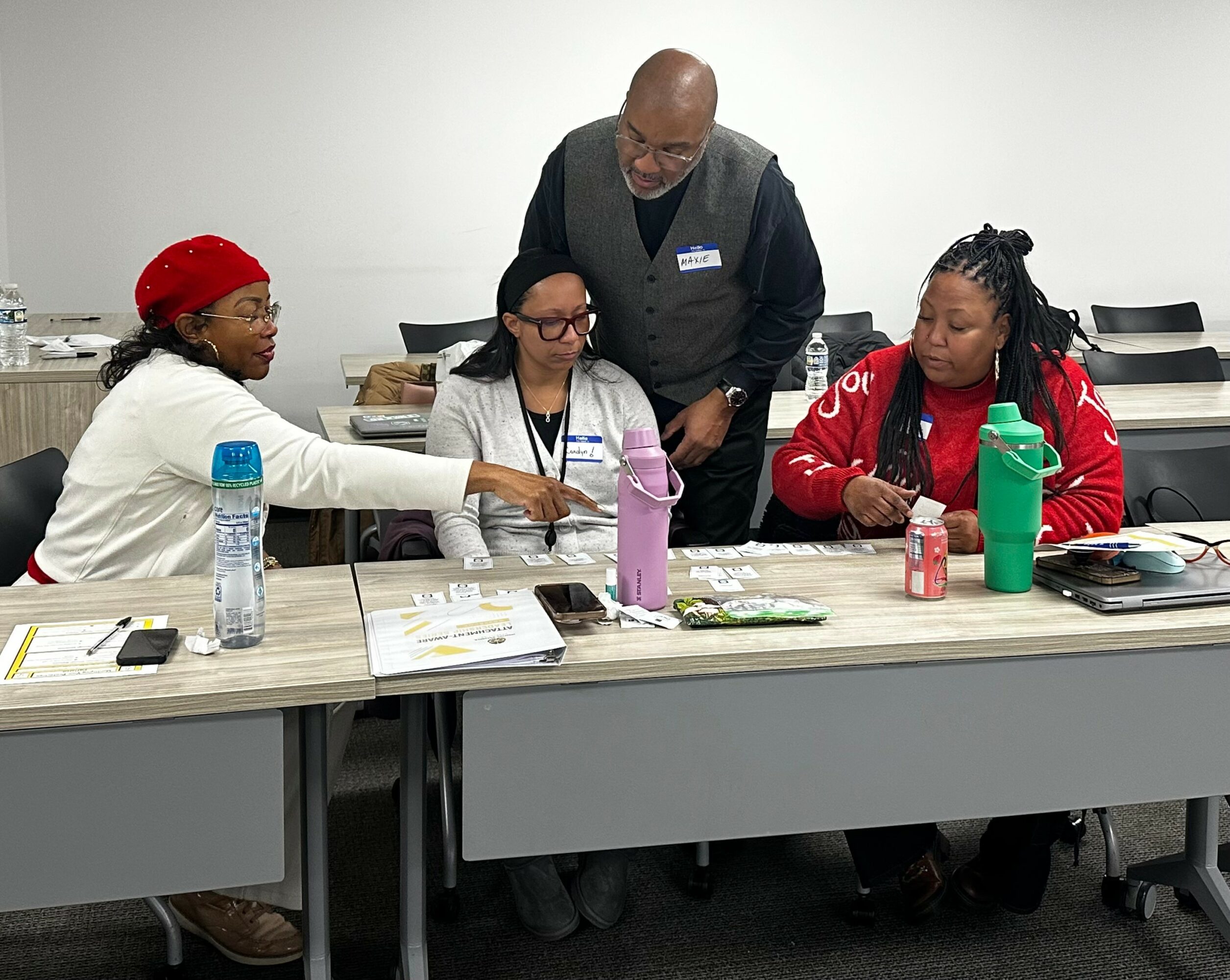 Four people sit at a table with water bottles, papers, and containers, engaging in a discussion or activity.