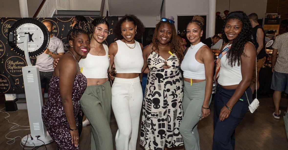 Group of seven women smiling and posing together indoors, some wearing white tops and pants, with a clock in the background.