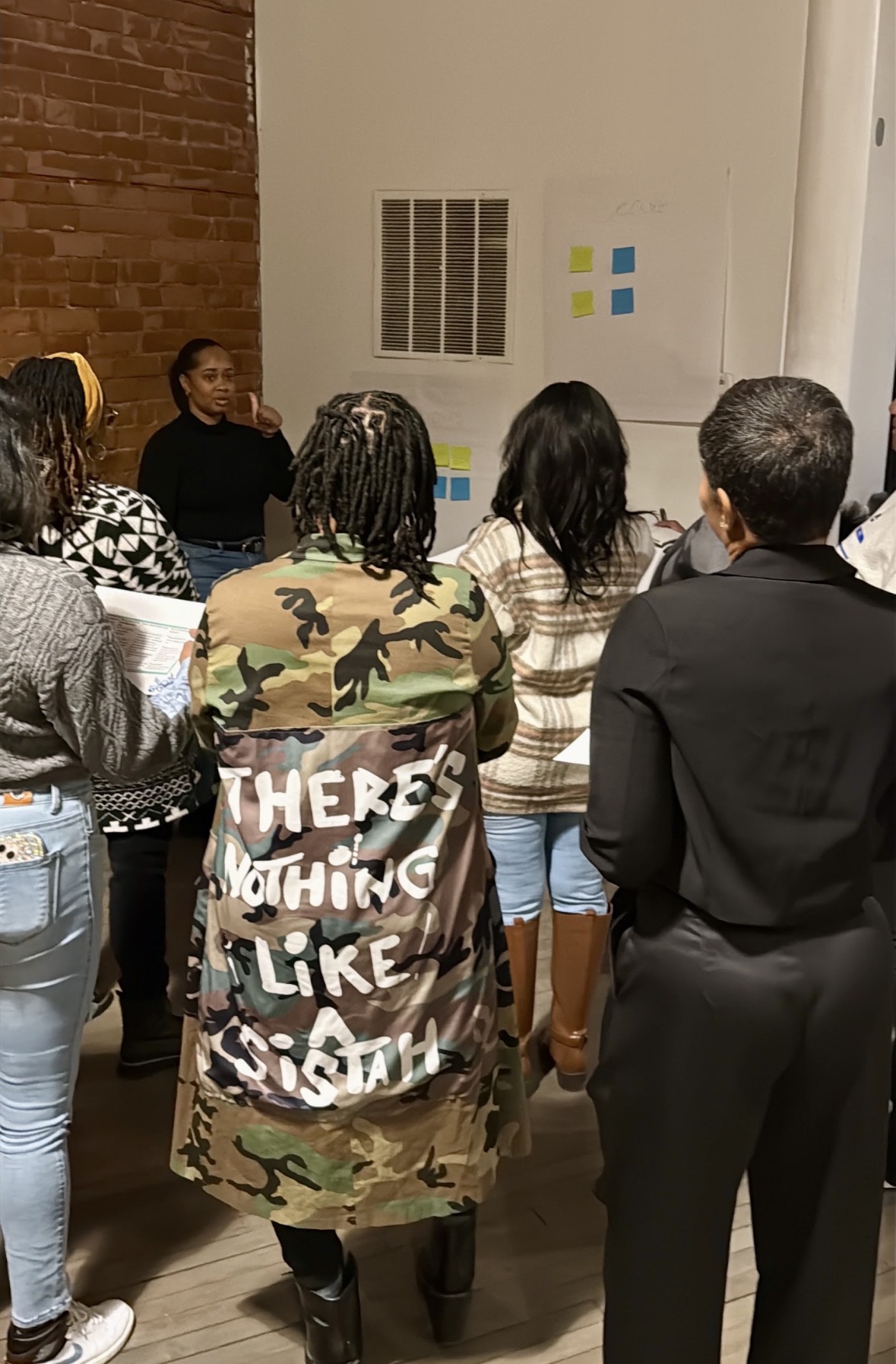 Group of people gathered indoors, facing a wall with charts or posters, listening to a speaker or presentation.