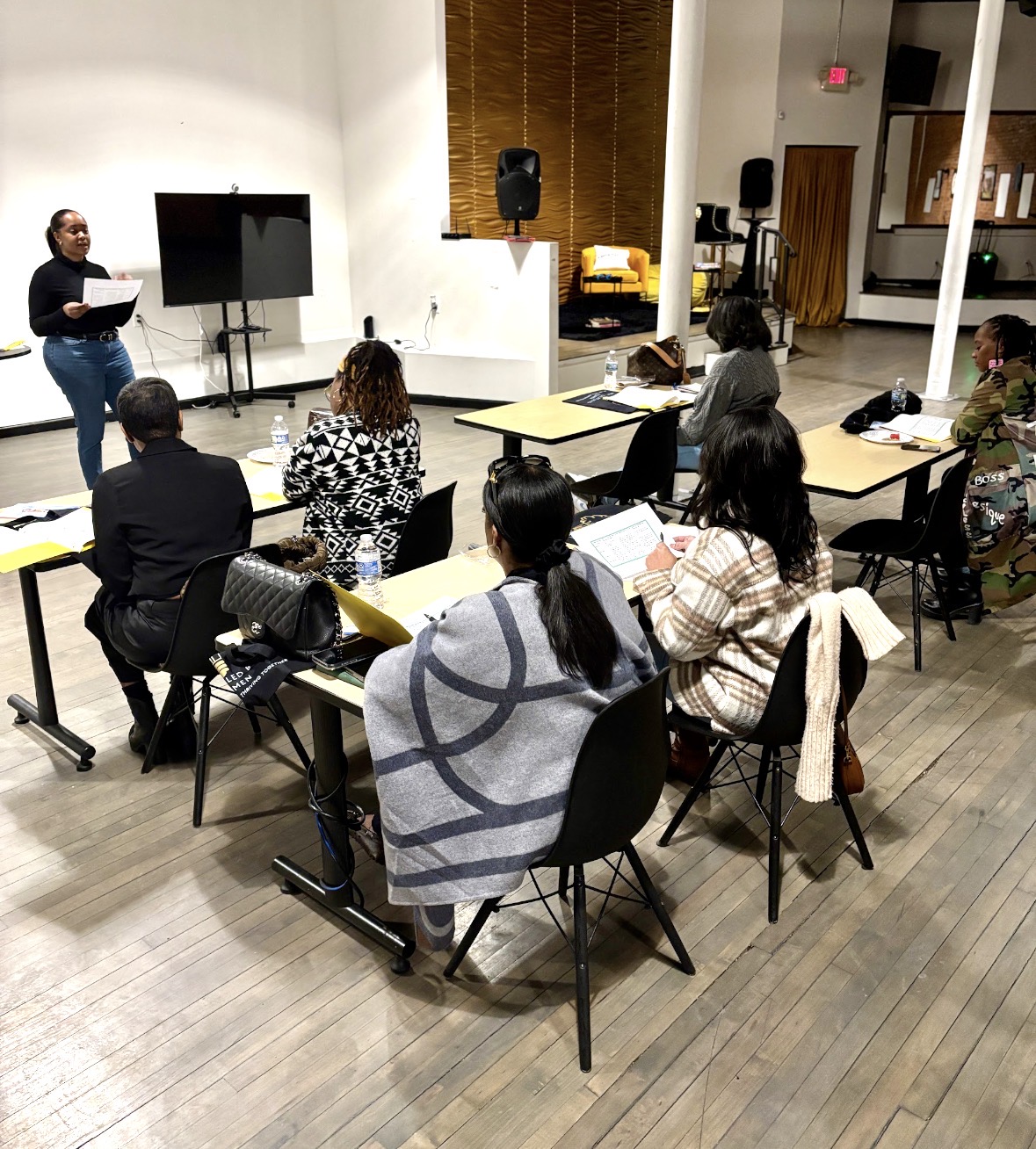 Person standing and speaking to seated group in a room with presentation equipment and shelves.