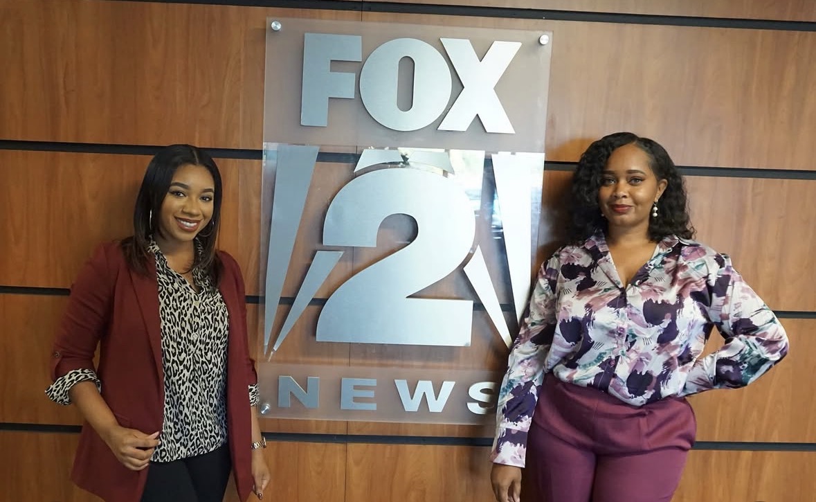 Two women standing in front of a FOX 21 News sign, smiling, with a wooden wall background.