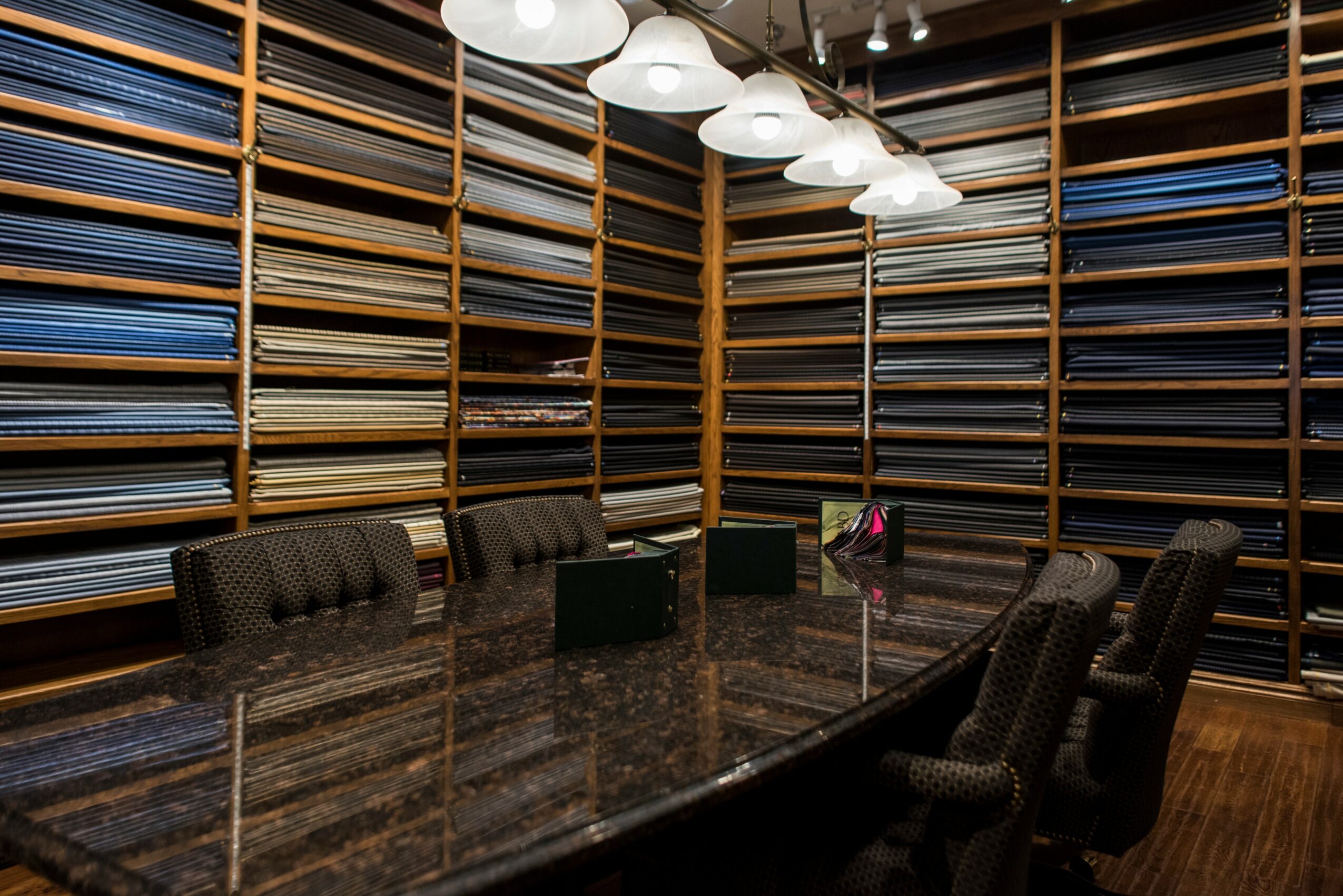 Room with shelves filled with fabric or material samples, a large table, and chairs, illuminated by ceiling lights.