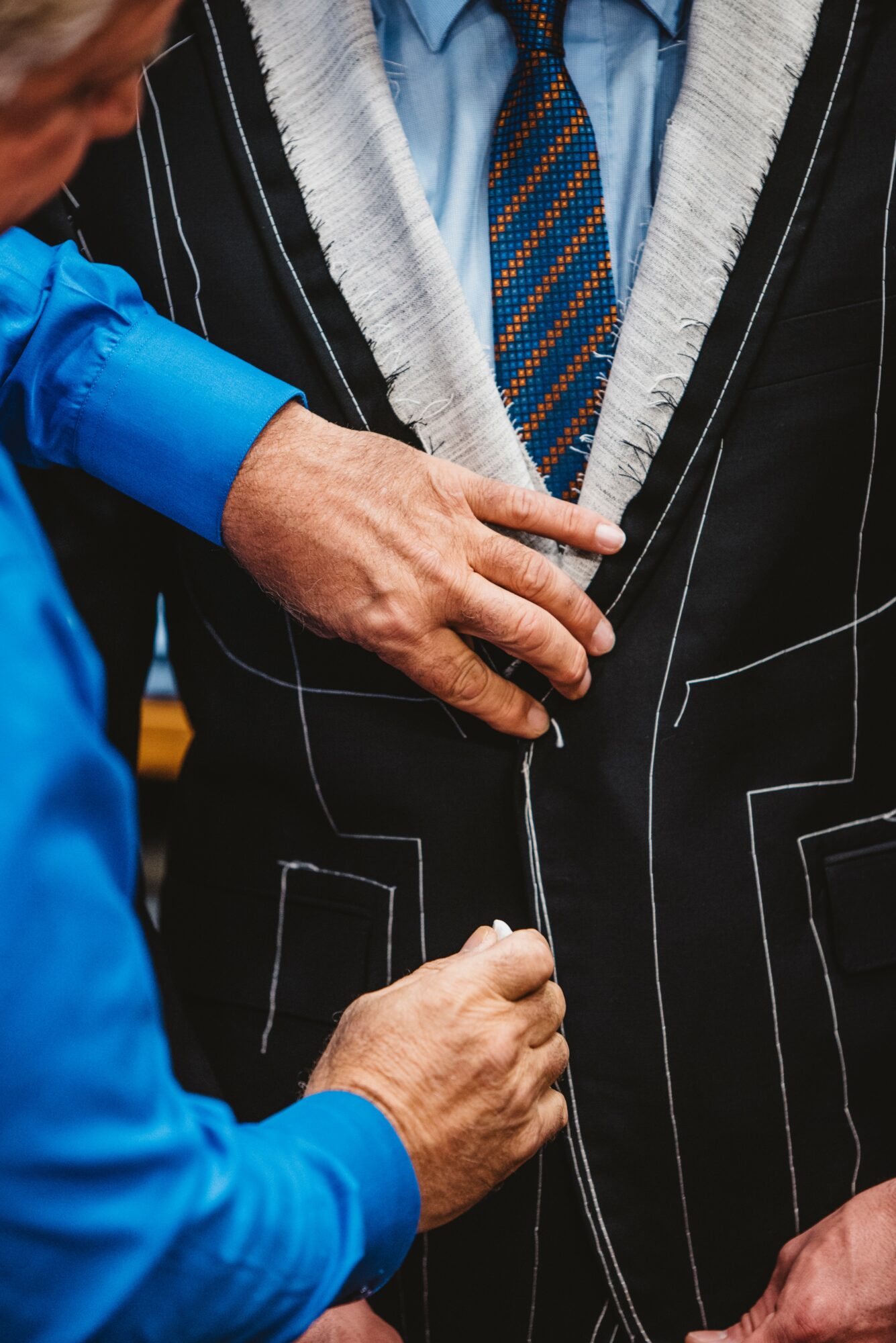 Two people, one in a blue shirt, are buttoning a black pinstripe suit jacket with white lines, close-up view.