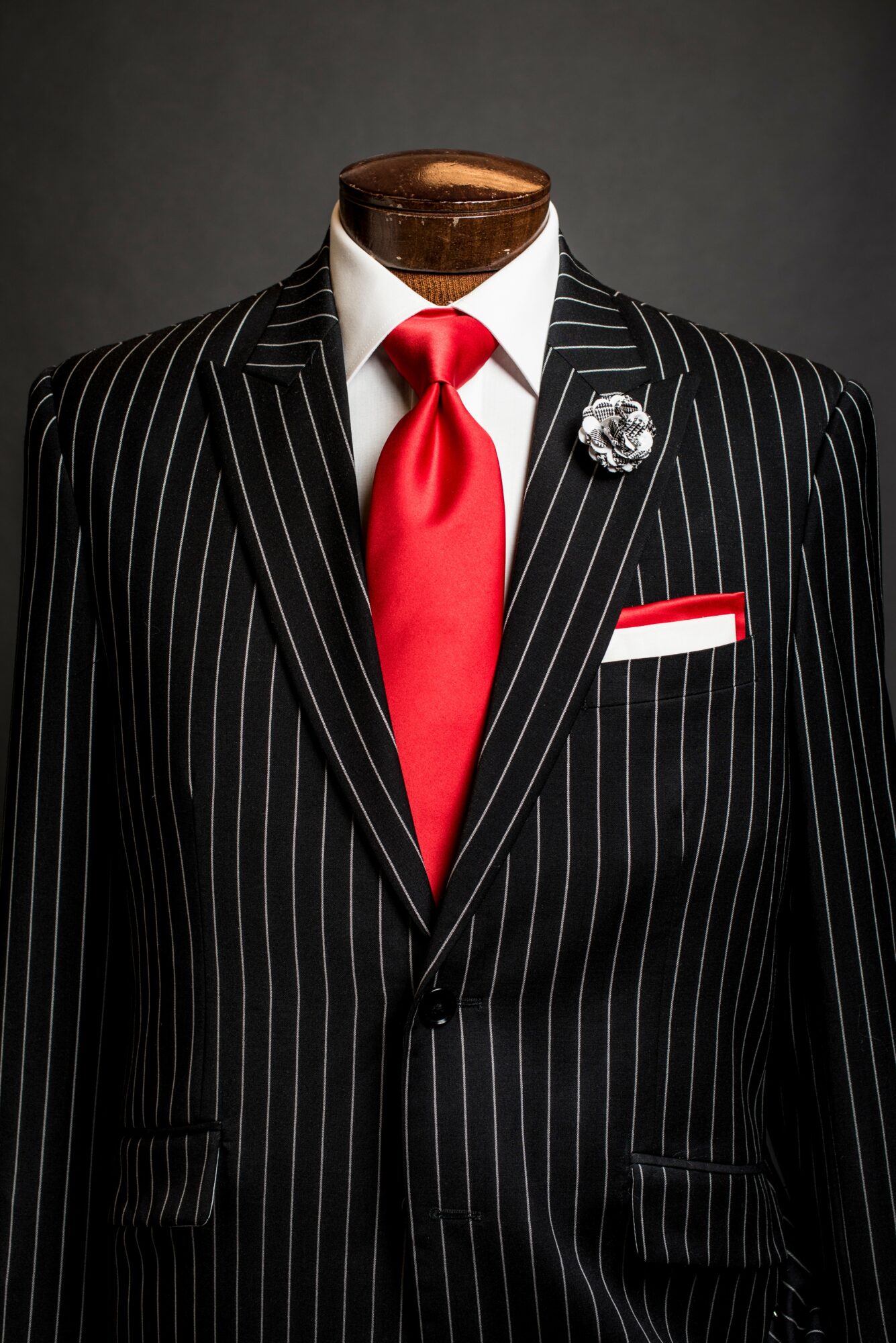 Mannequin dressed in a black pinstripe suit with a red tie, white shirt, and decorative pin, against a dark background.