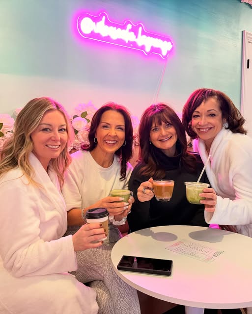 Four women sitting at a table in a cafe, smiling and holding drinks, with a pink neon sign on the wall behind them.