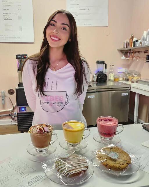Young woman with long dark hair smiling behind a counter with three ice cream sundaes and cookies.