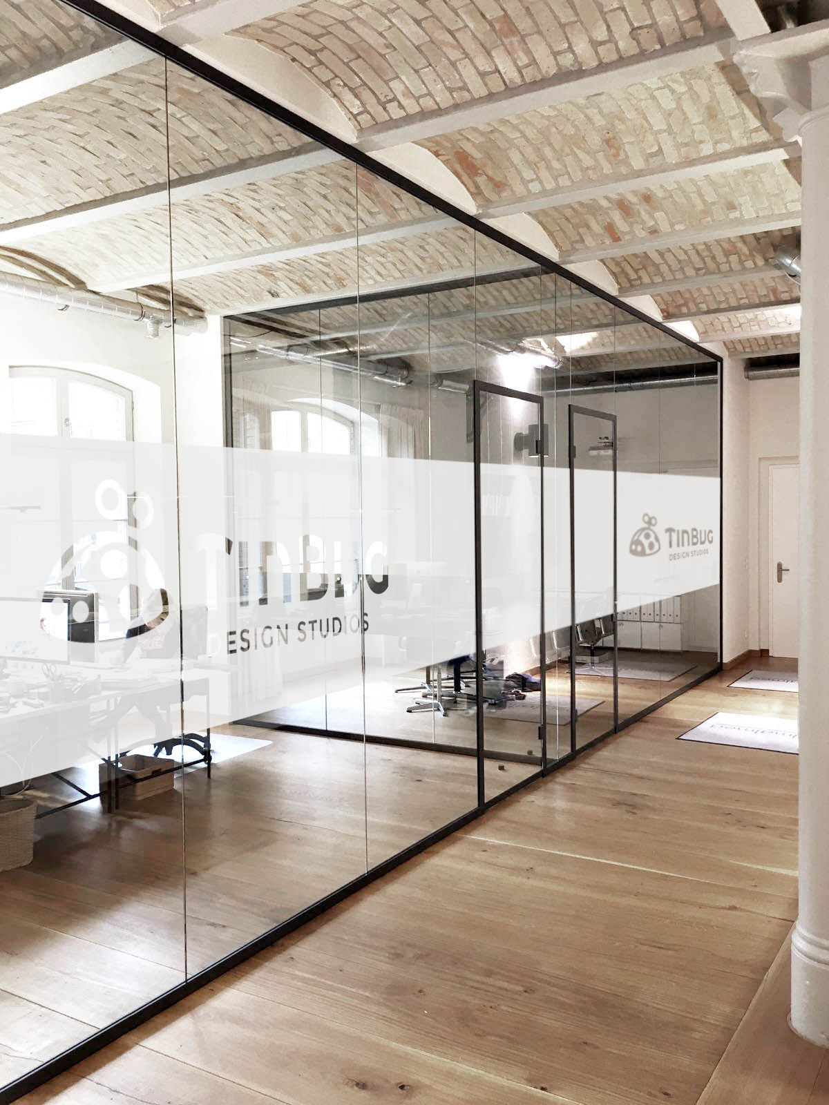 Glass-walled office with branding, brick ceiling, wooden floor, and natural light, reflecting interior space.
