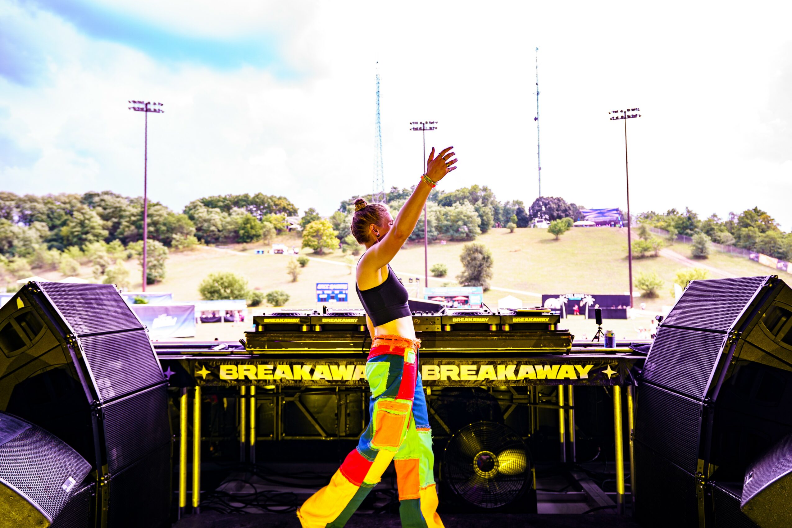 Person on stage with arms raised, wearing colorful pants, in front of speakers and a bright outdoor background.