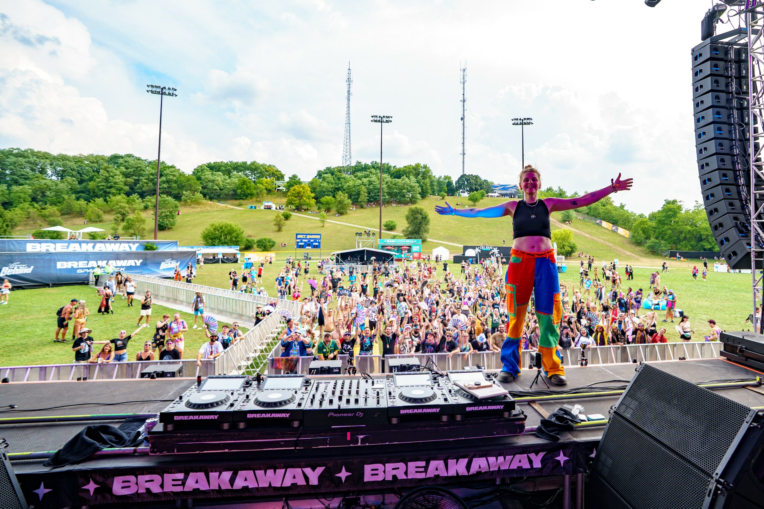 DJ on stage with a crowd at outdoor music festival, green hill, blue sky, and tall speakers visible.