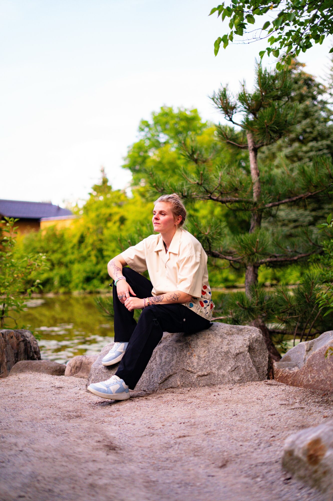 Woman sitting on a large rock outdoors, surrounded by trees and greenery, with a building in the background.