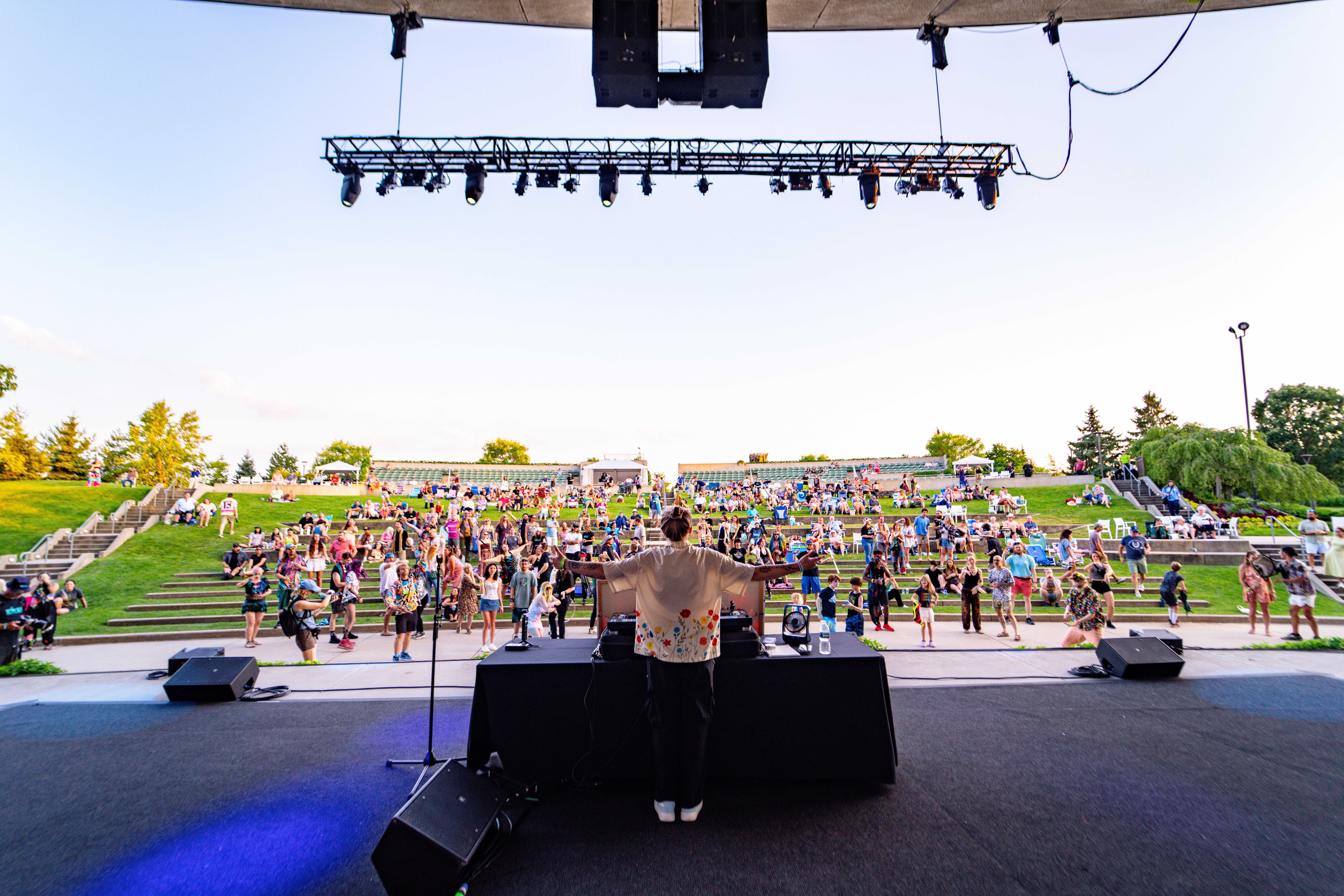 Performer on stage facing an outdoor audience with tiered seating, stage equipment visible, and a clear sky.