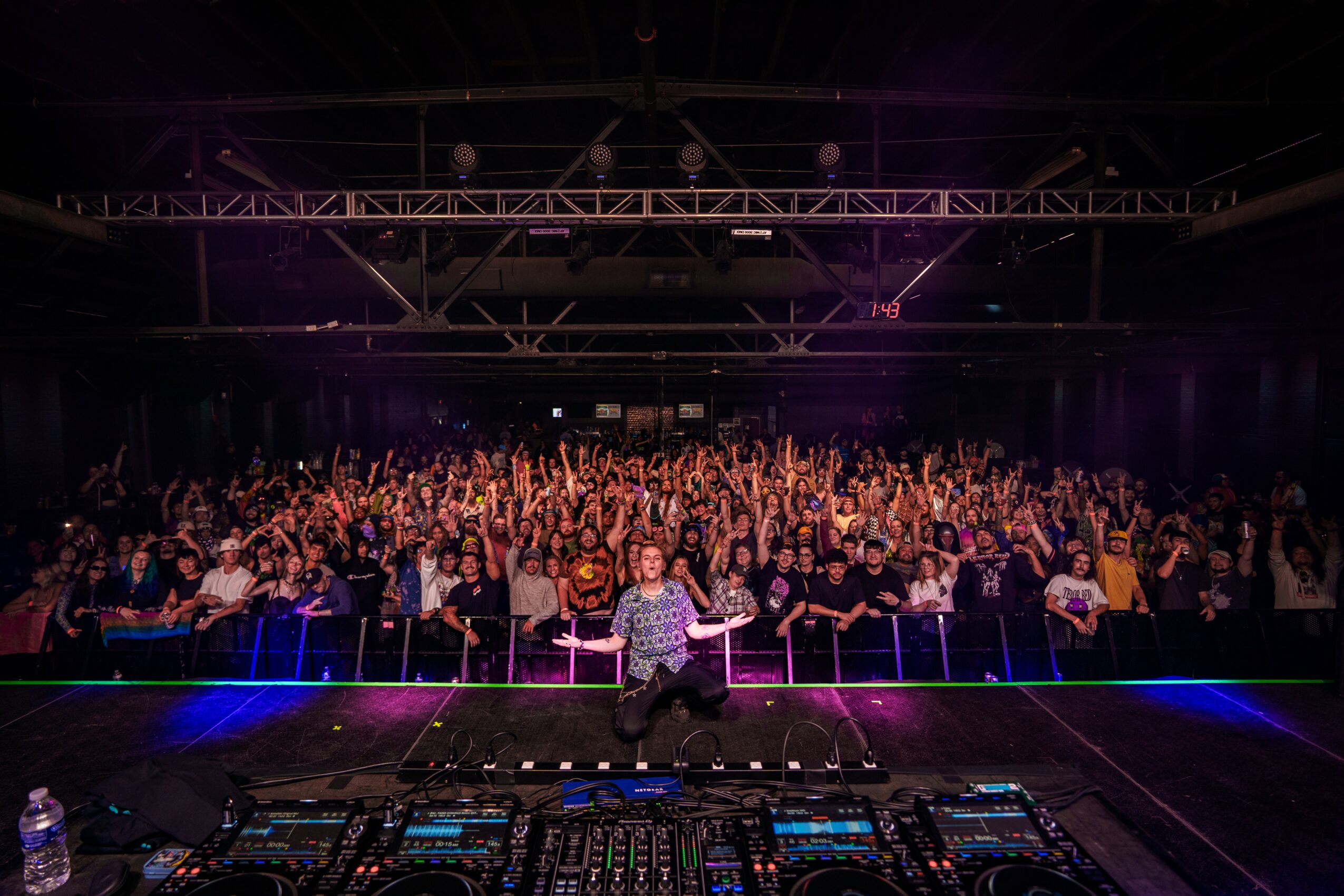 DJ performing on stage with a large crowd in a dark indoor venue, colorful lights, and equipment in front, 20 words.
