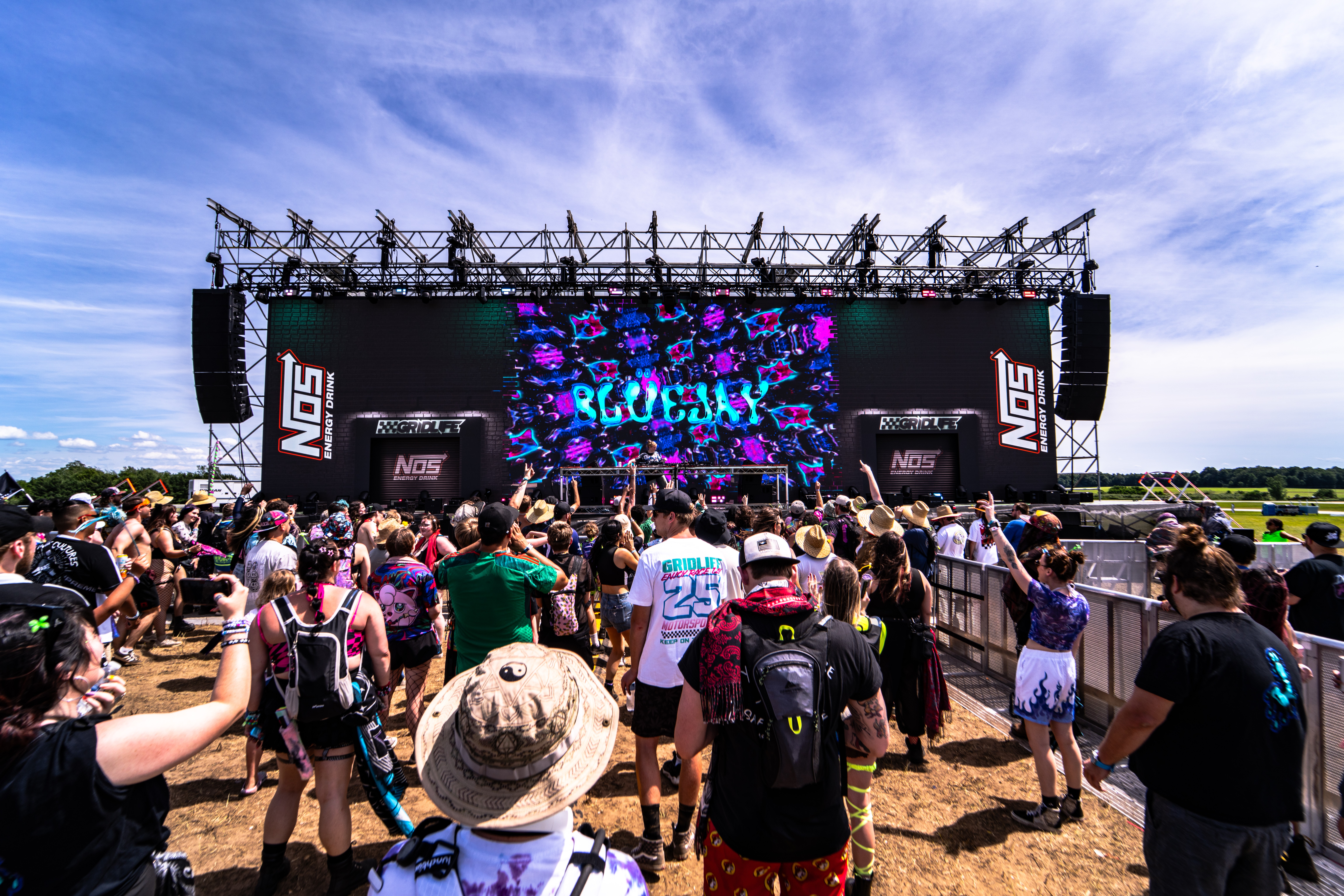 Crowd of people watching an outdoor concert stage with a large screen and speakers, under a partly cloudy sky.