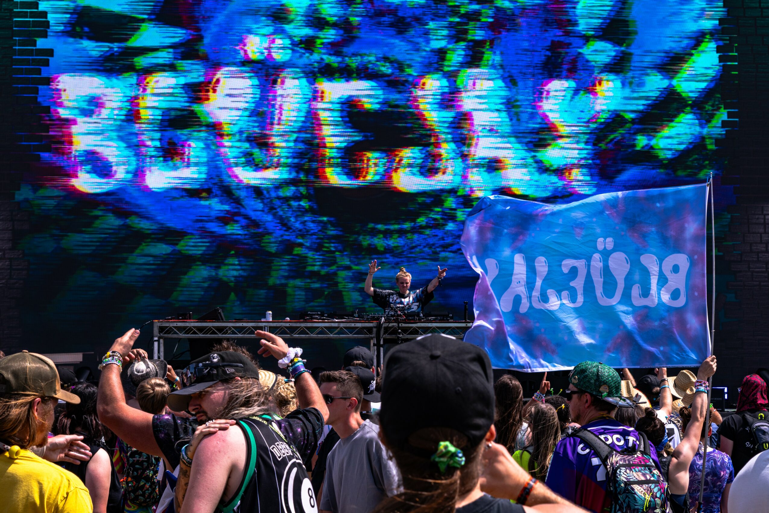 Crowd at a music festival with a large digital screen displaying colorful text and a performer on stage, some people taking photos.