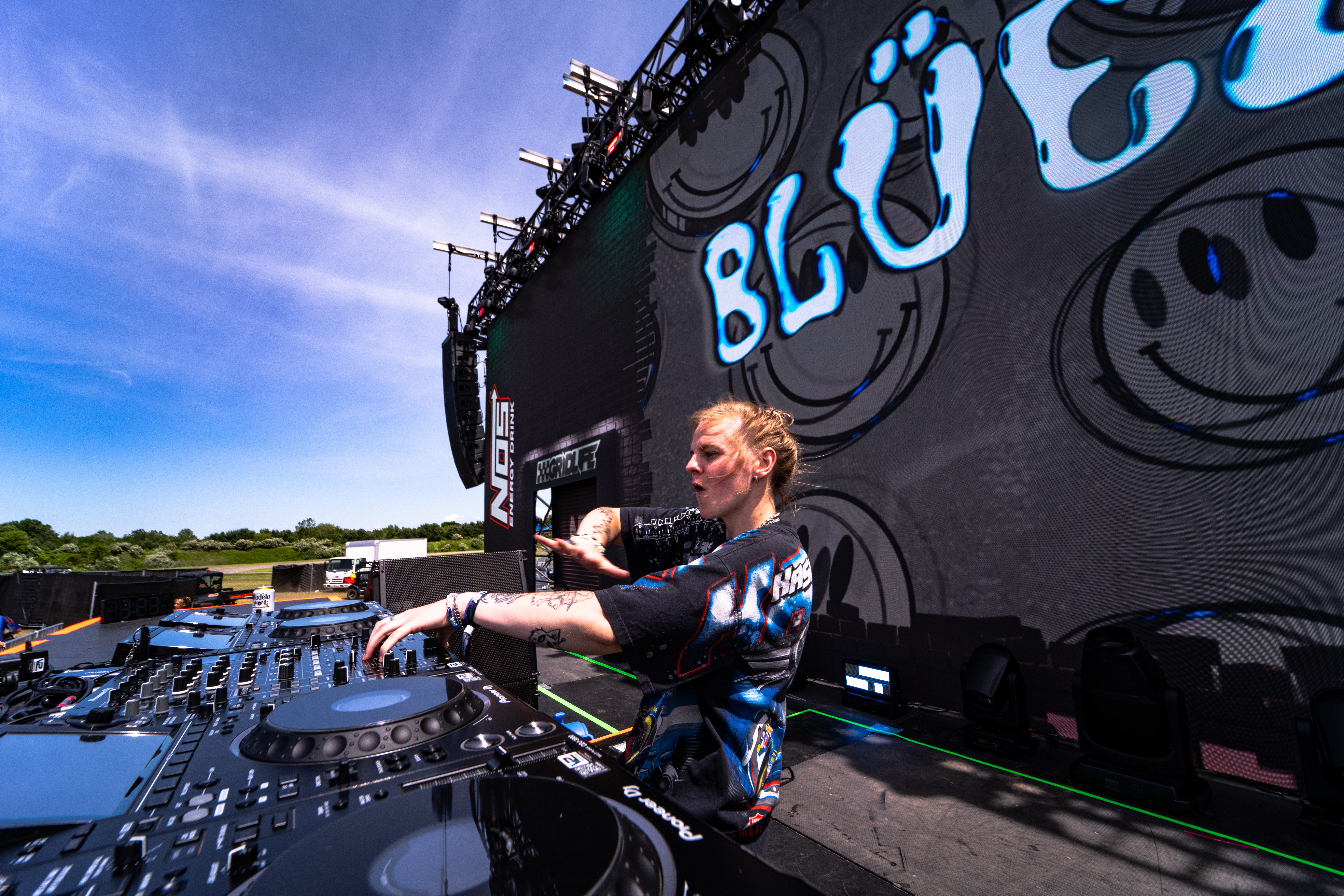 Female DJ performing on stage with DJ equipment, large black backdrop with blue and white graffiti-style text, outdoor setting, clear sky