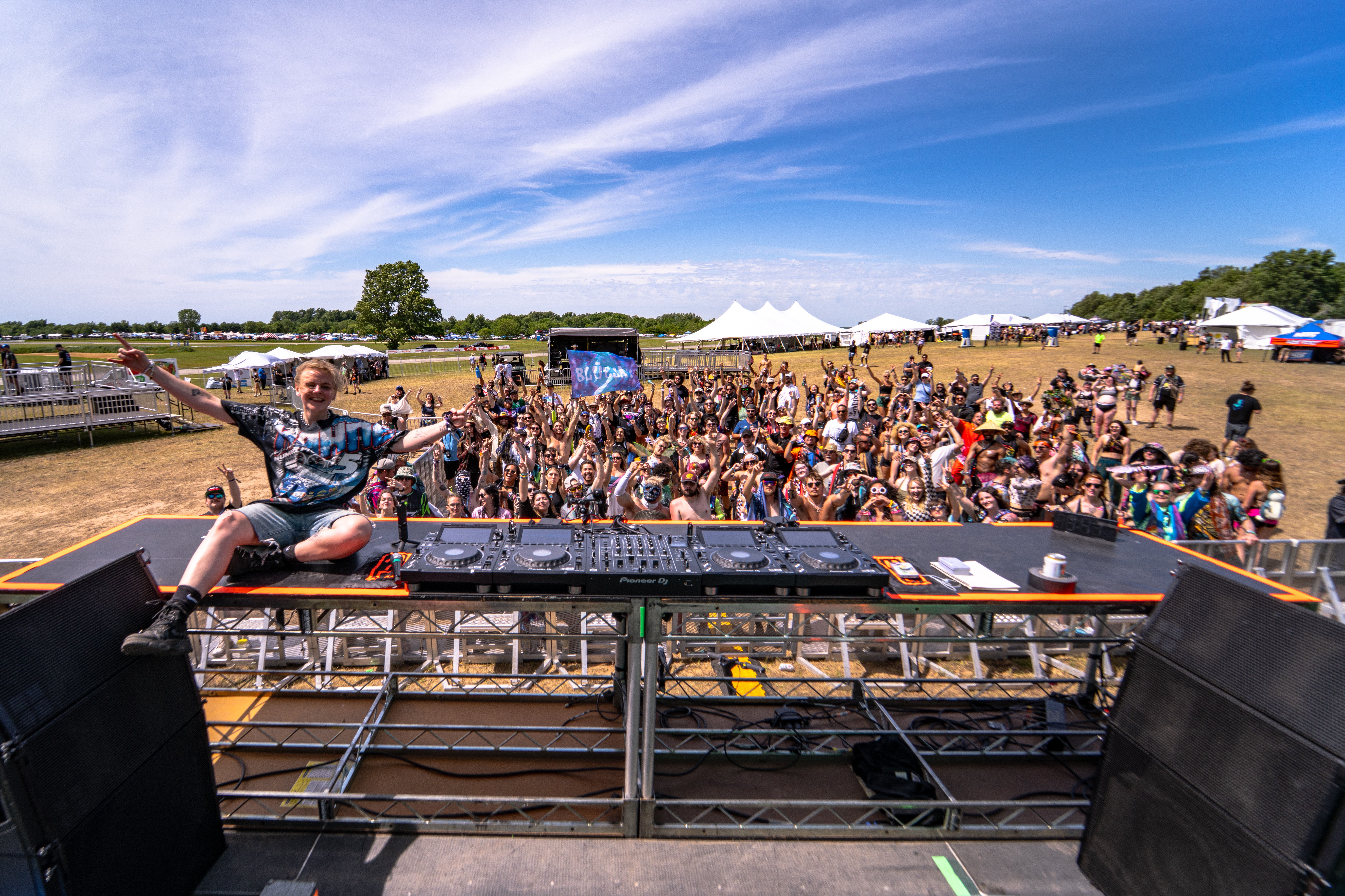 Person with arms raised on stage overlooking a large outdoor crowd at a festival under a blue sky.