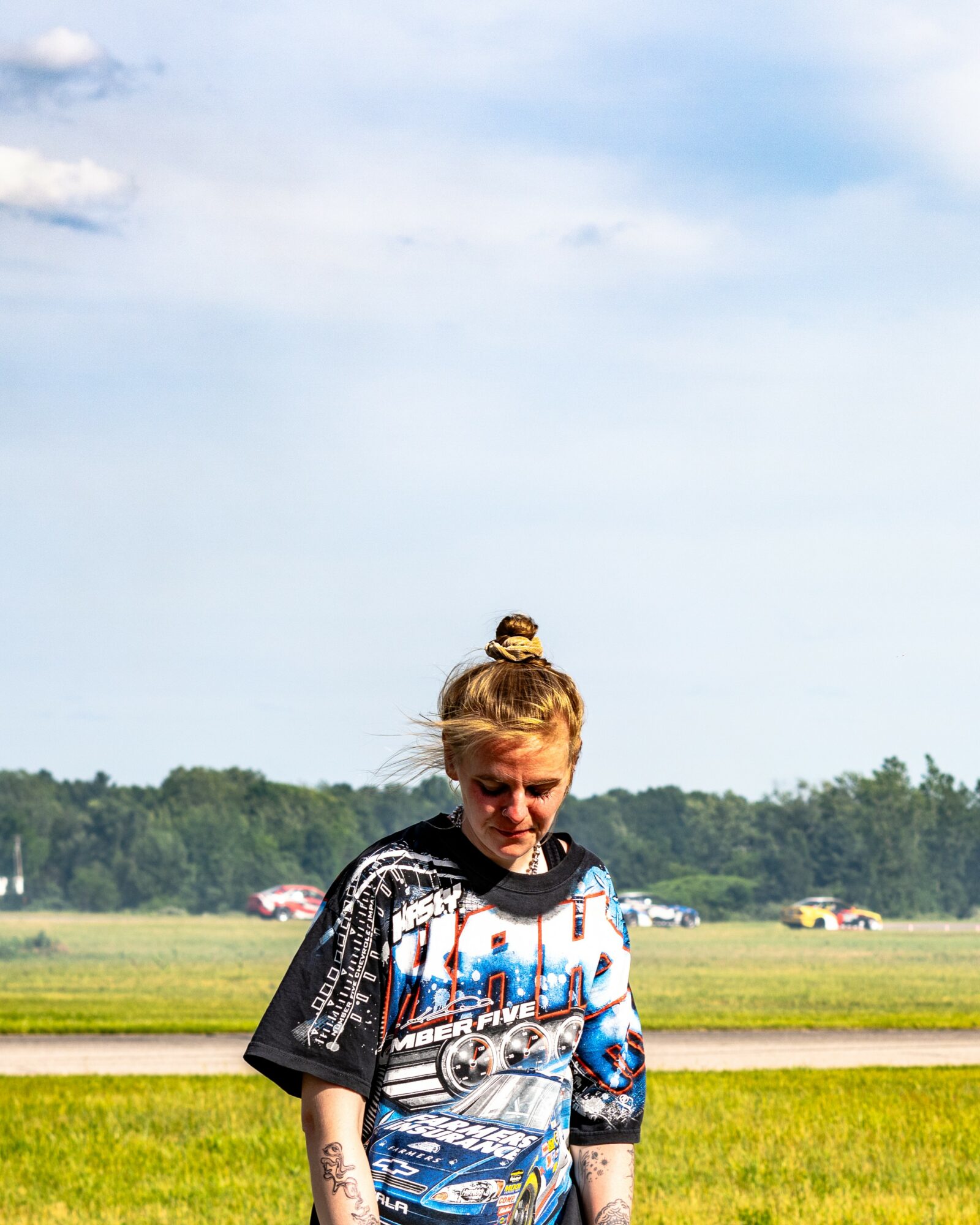 Girl with a topknot hairstyle standing outdoors in a grassy field, looking downward, with a blue sky and trees in the background.