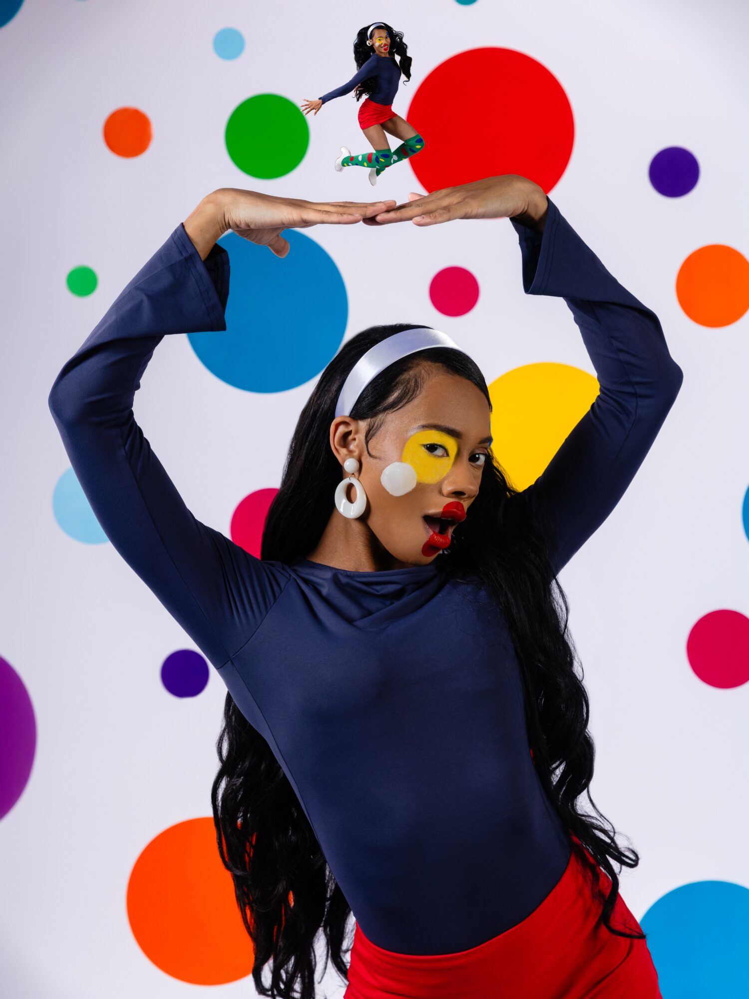Woman with long dark hair and colorful makeup balances a tiny woman on her hands against a polka dot background.