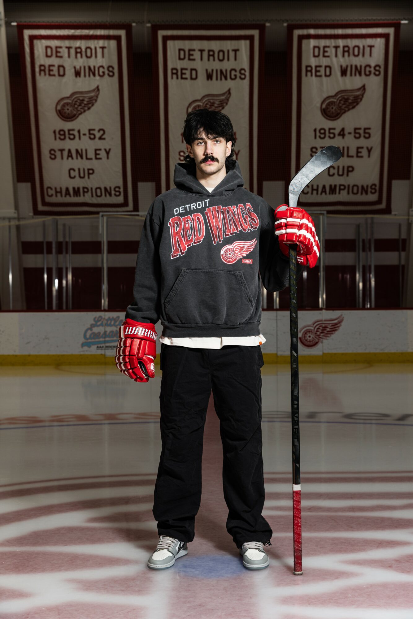 Person standing on ice rink holding hockey stick, wearing Detroit Red Wings hoodie and gloves, with banners in background.