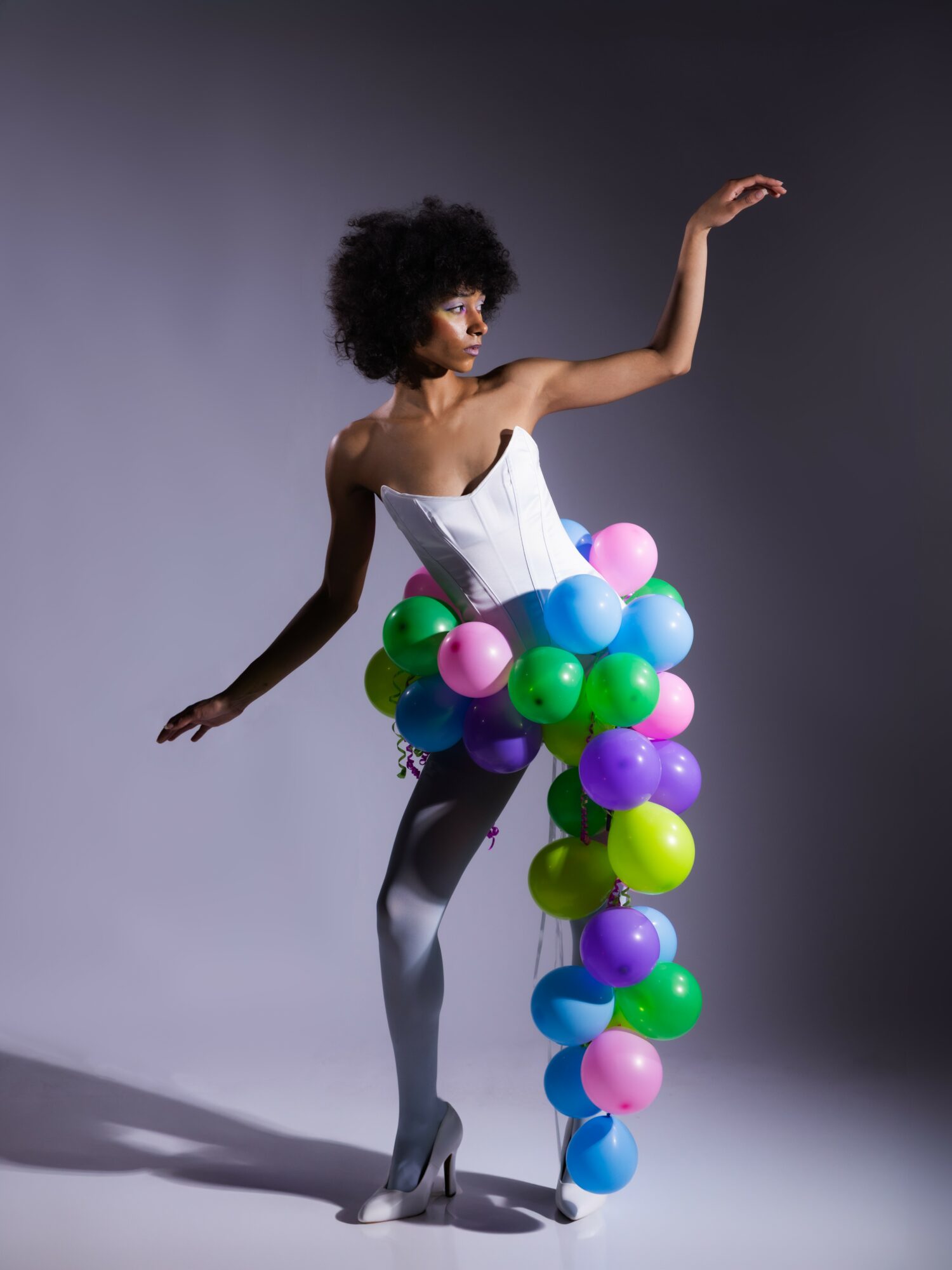 A woman with curly hair in a white top and gray leggings stands with one arm raised, surrounded by colorful balloons.