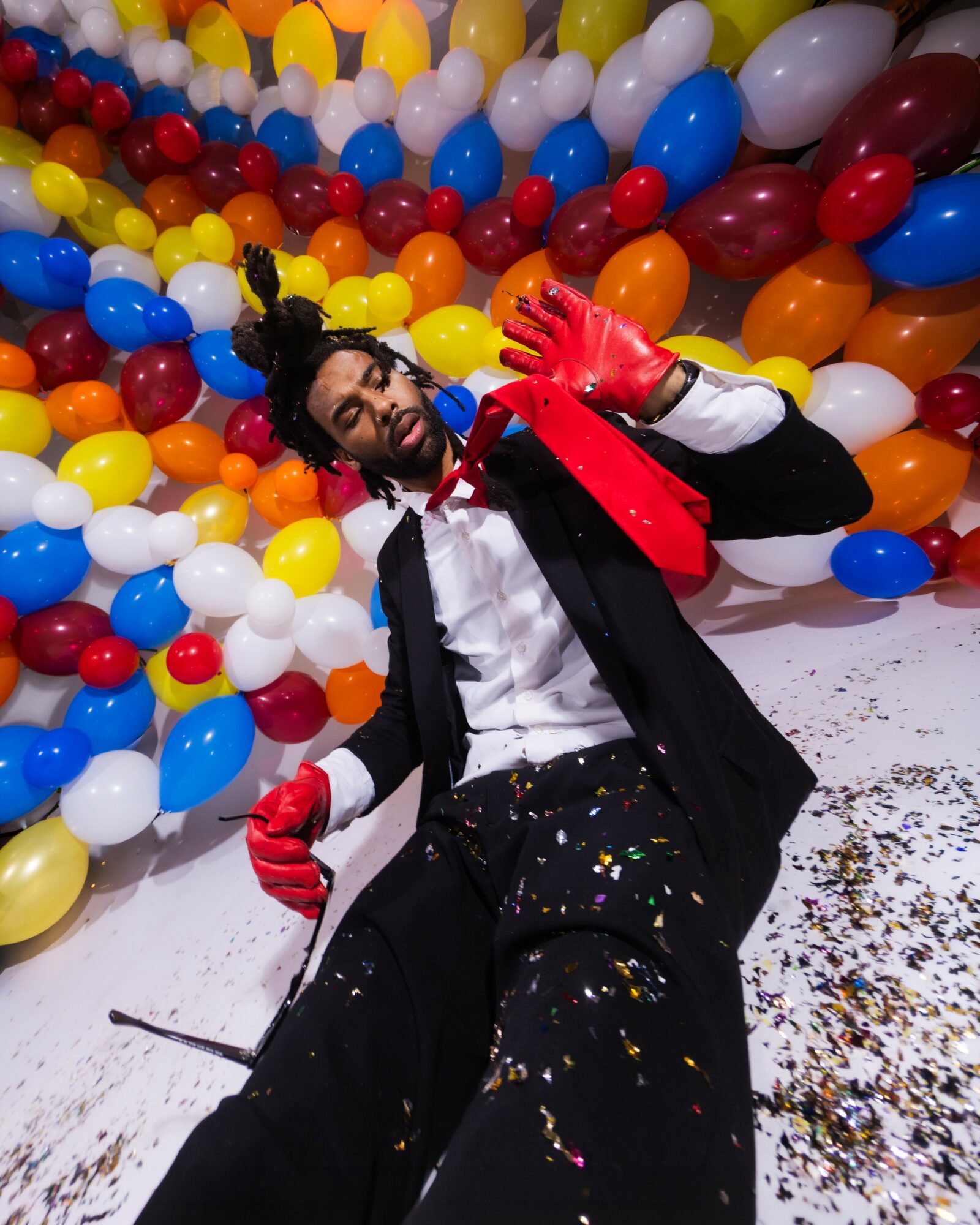 Person lying on the floor with colorful balloons background, wearing a black coat, white shirt, red gloves, and red tie, smiling.
