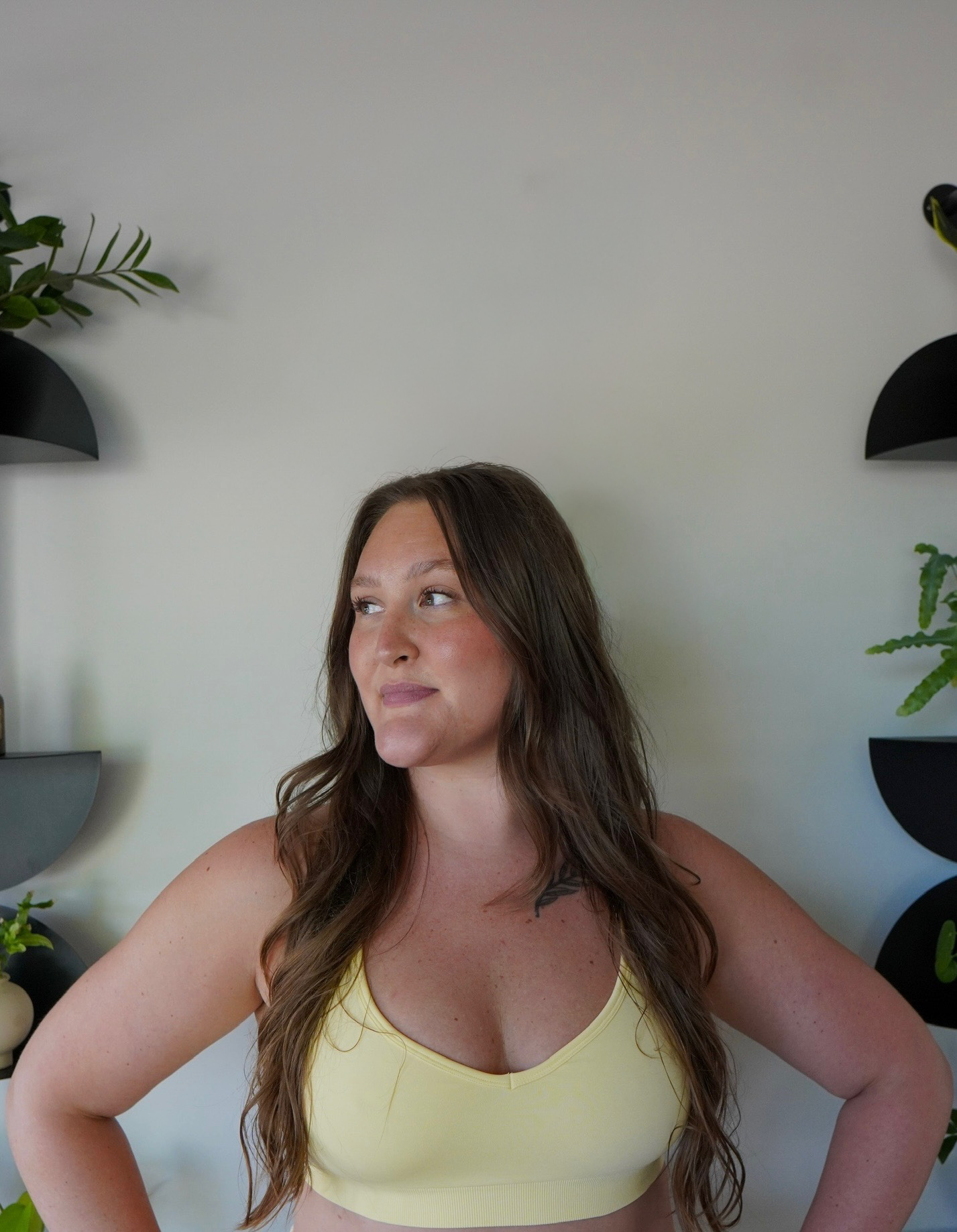 Woman with long brown hair wearing a yellow top, standing indoors with plants on the wall behind her.