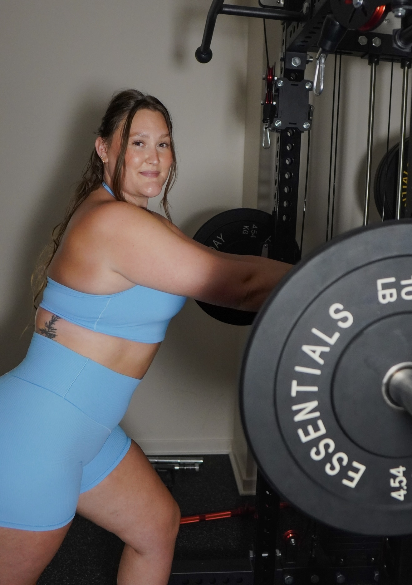 Woman in blue workout outfit lifting a barbell in a gym, smiling at the camera.