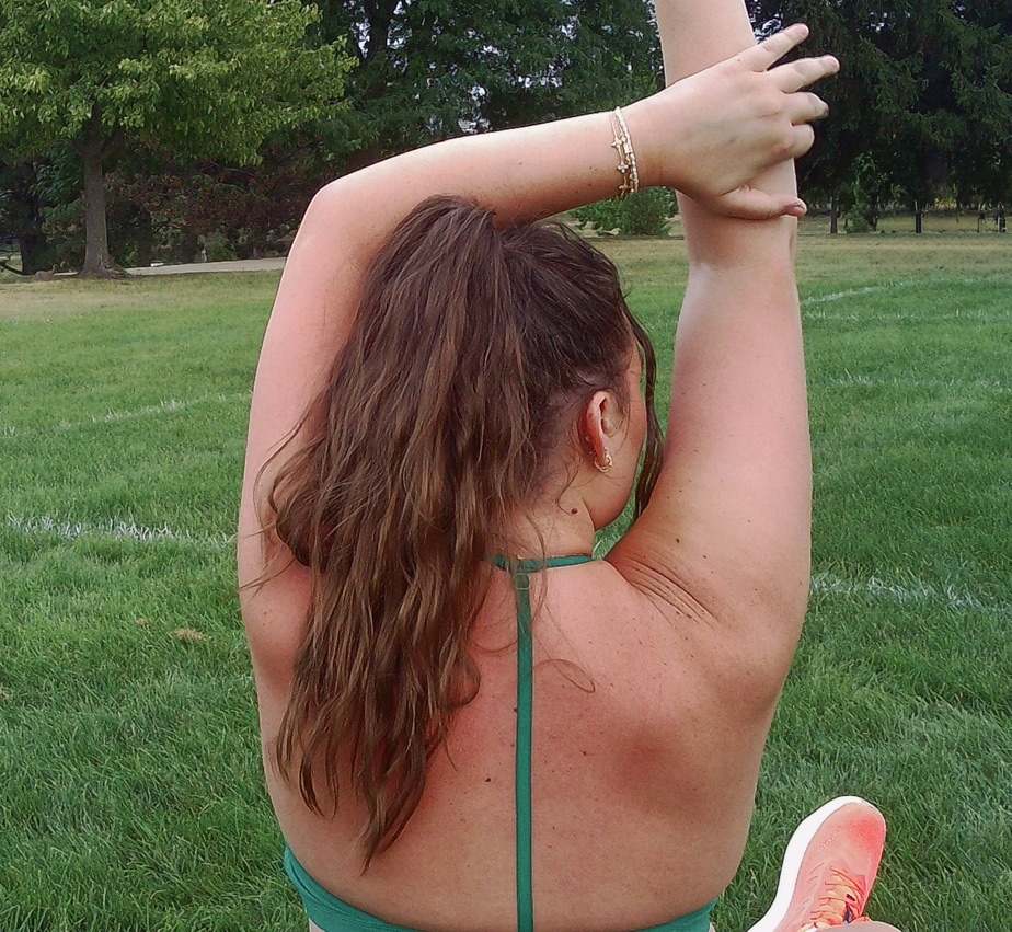 Person with long hair stretching arms above head outdoors on grass, back facing camera, wearing a green top.