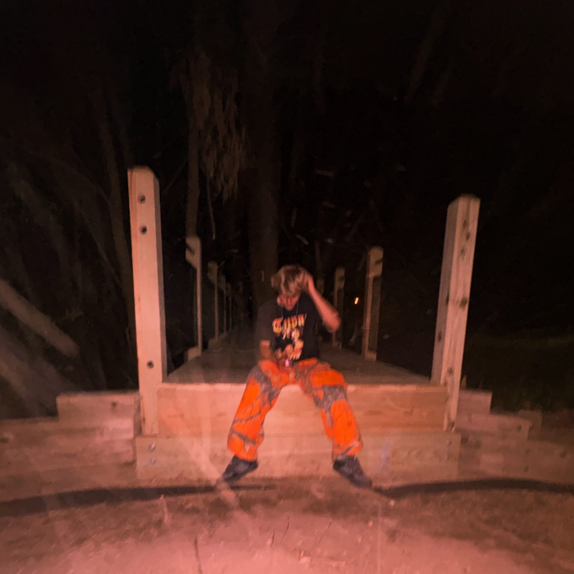 Person in orange pants and a black shirt standing on a wooden platform at night, holding a helmet, with dark background.