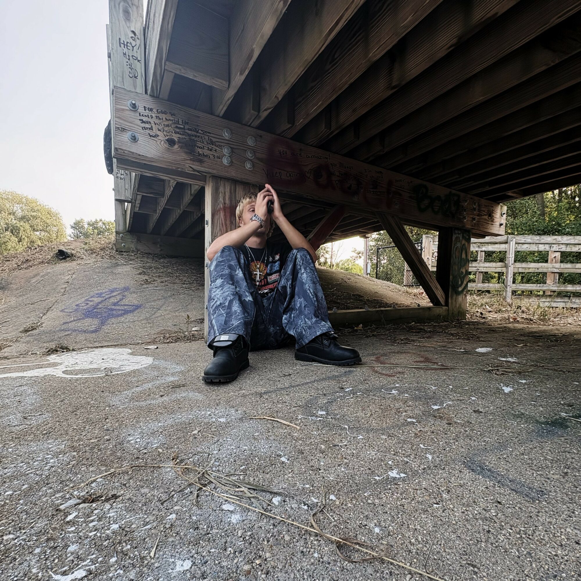 Person sitting under a bridge with hands covering face, wearing baggy jeans and black boots, on a dirt ground.