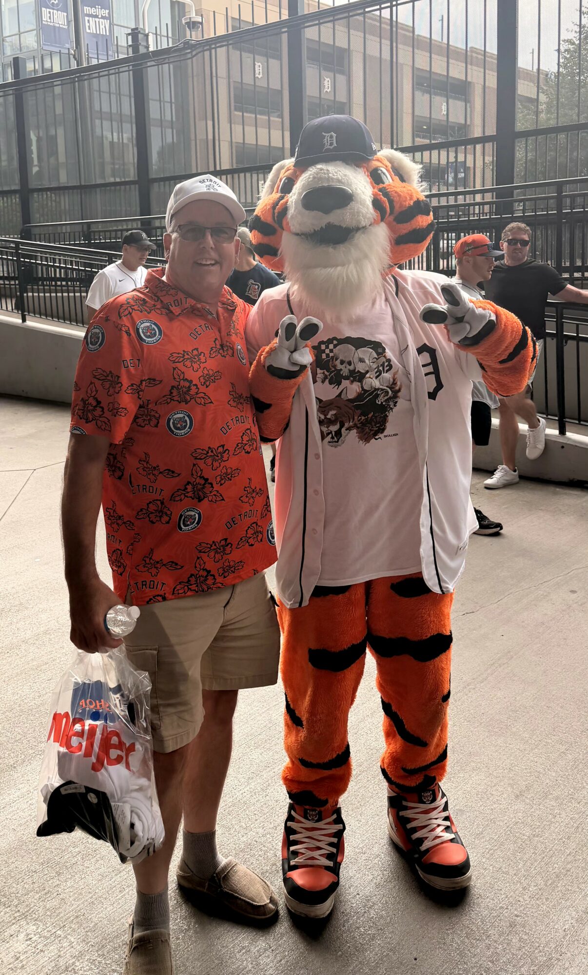Person in a red shirt and shorts standing next to a tiger mascot in a sports jersey, with people in the background.