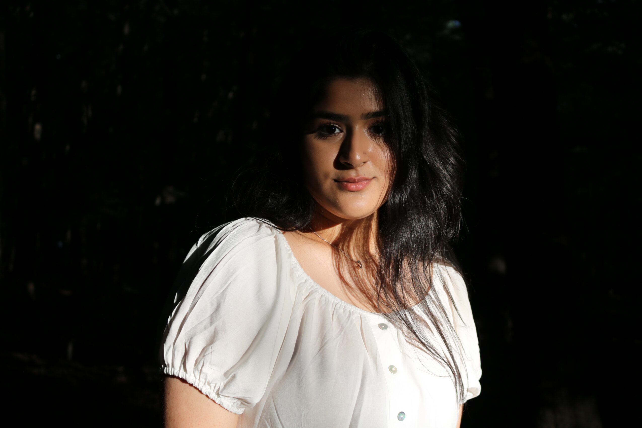 Young woman with dark, wavy hair wearing a white blouse, standing outdoors at night.