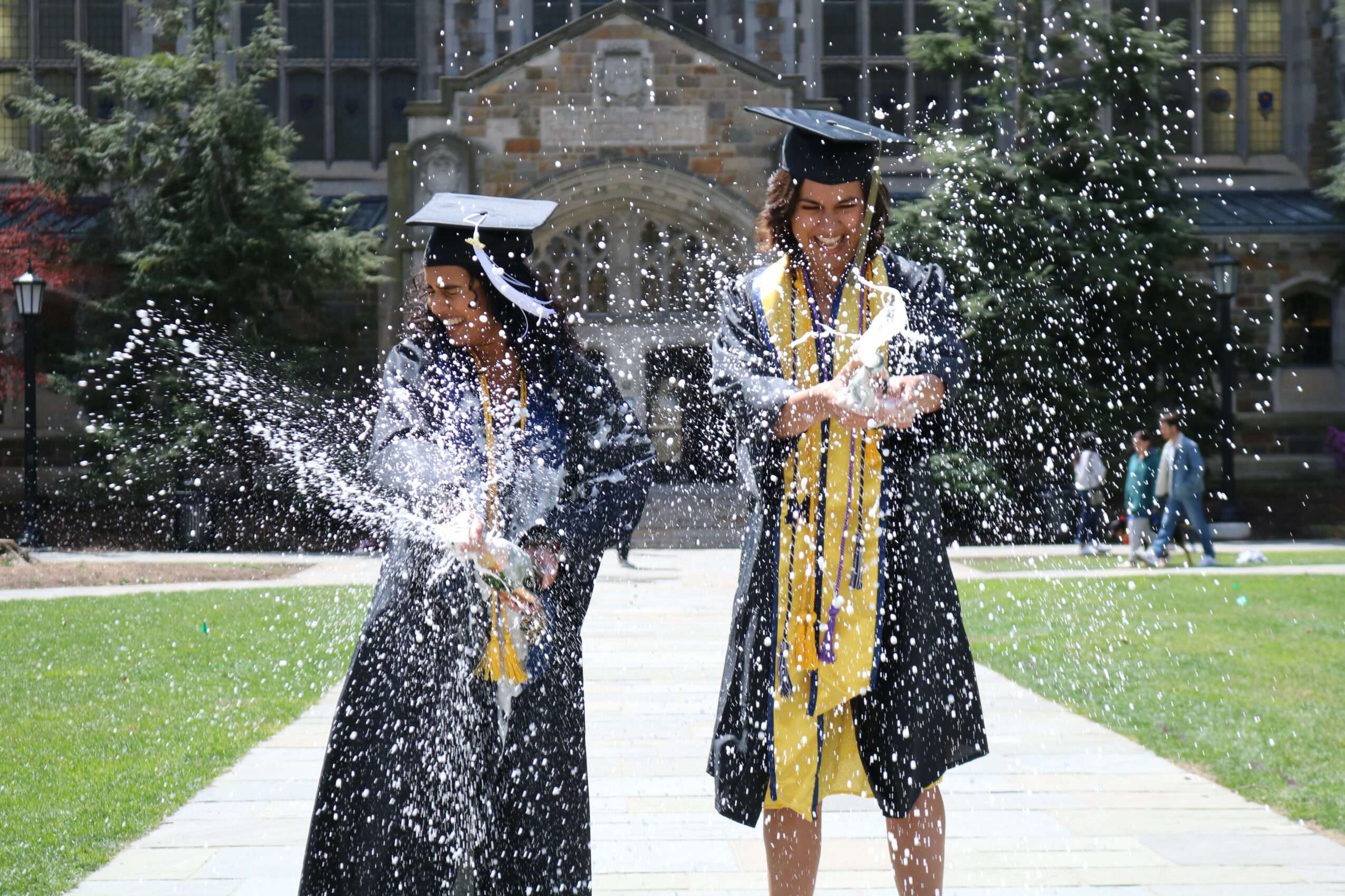 Two graduates in caps and gowns celebrate outdoors, throwing water on each other on a sunny day.