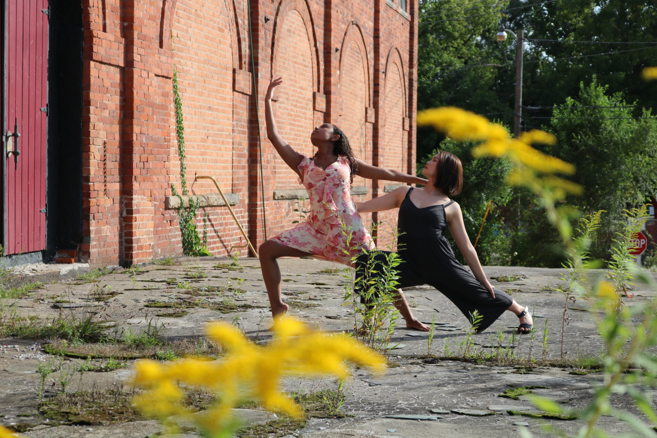 Two women sit on a stone ledge in front of a brick wall, one stretching and the other sitting back, with yellow flowers in foreground